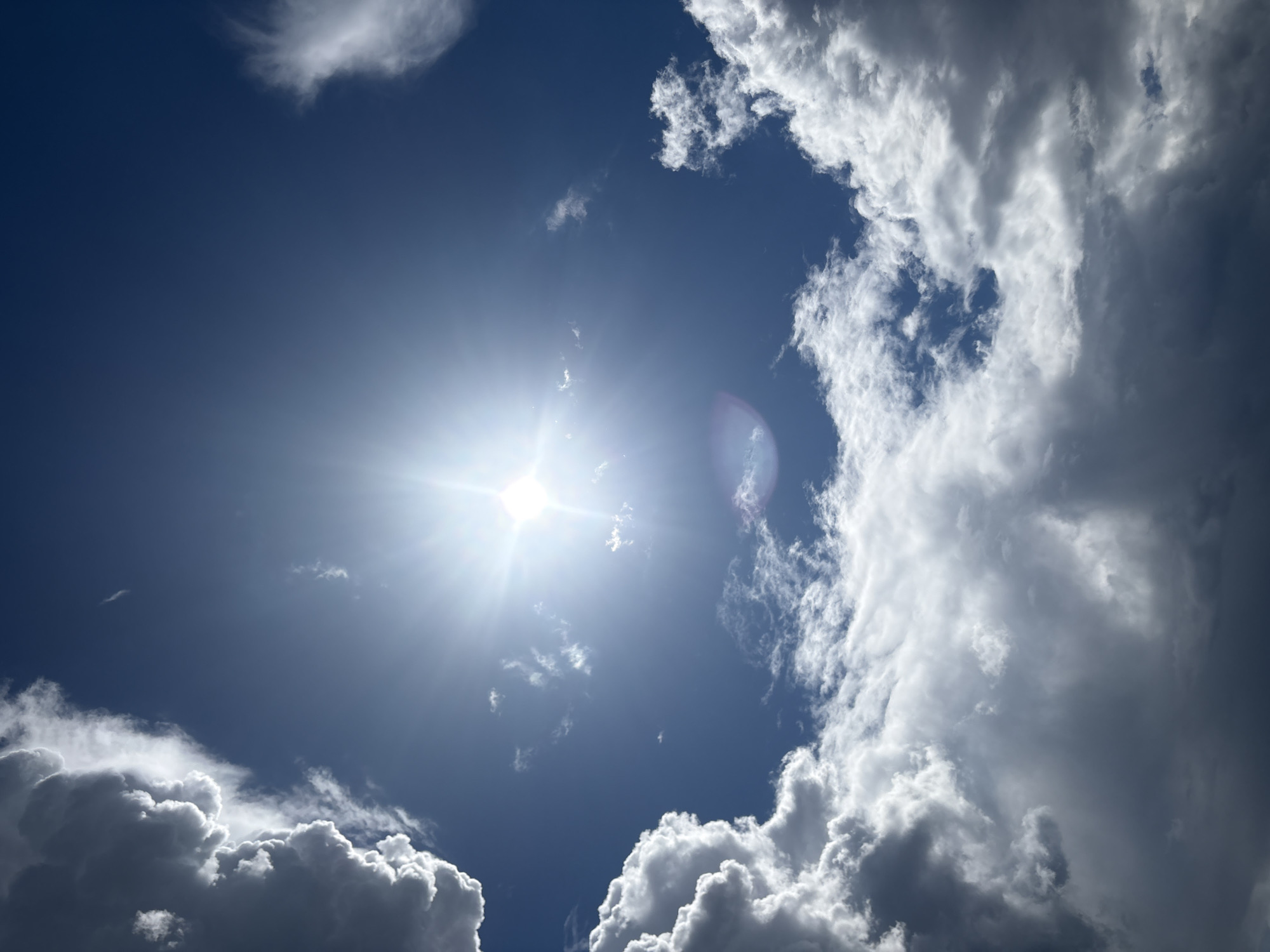 A wide-angle, low-angle shot captures a bright sun shining through a deep blue sky. The sun is the dominant subject, emitting brilliant rays that create a lens flare effect on the right side of the frame. Scattered clouds populate the sky, with larger, cumulus formations framing the scene on the left and right edges. These clouds range in color from pure white where illuminated by the sun to darker, grayer tones in shadowed areas, suggesting depth and varying densities. Delicate wisps of cirrus clouds are also visible, further enhancing the dynamic nature of the sky. The overall impression is one of a bright, possibly warm day, with a mix of clear sky and cloud cover. The image conveys a sense of openness and natural beauty. The time of day appears to be midday due to the bright, high sun. No people, objects, or text are visible in the image. The location context is Orsk, Russia, but no specific landmarks or cues confirm this.