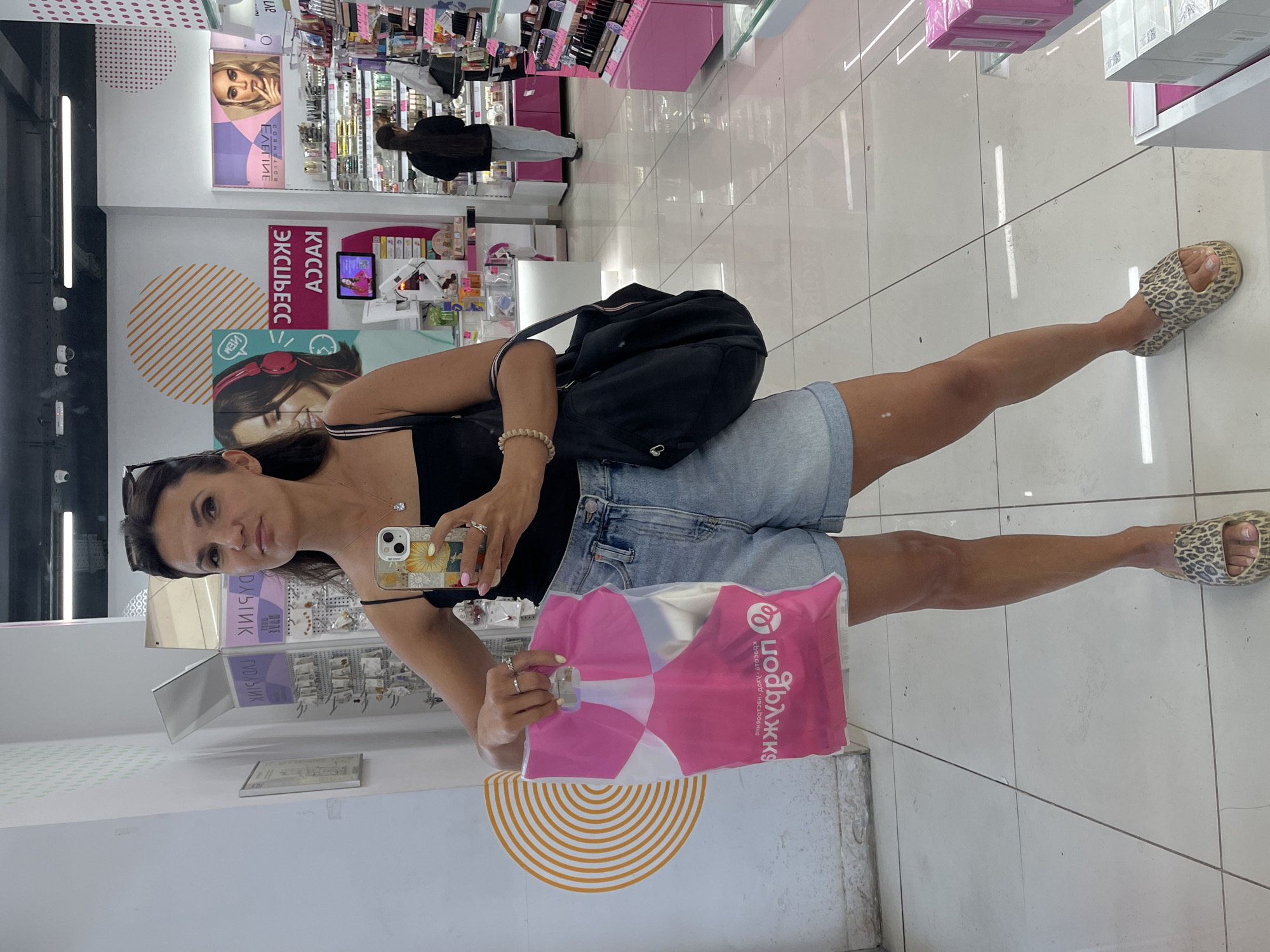 A woman is taking a selfie in a beauty store. She is wearing a black tank top, denim shorts, and leopard-print slides. She is holding a pink shopping bag in her left hand and her phone in her right hand. The store appears to be well-lit with white tiled floors and shelves stocked with makeup and beauty products. There is a woman with long dark hair in the background, also looking at products. The store is in Moscow, Russia.