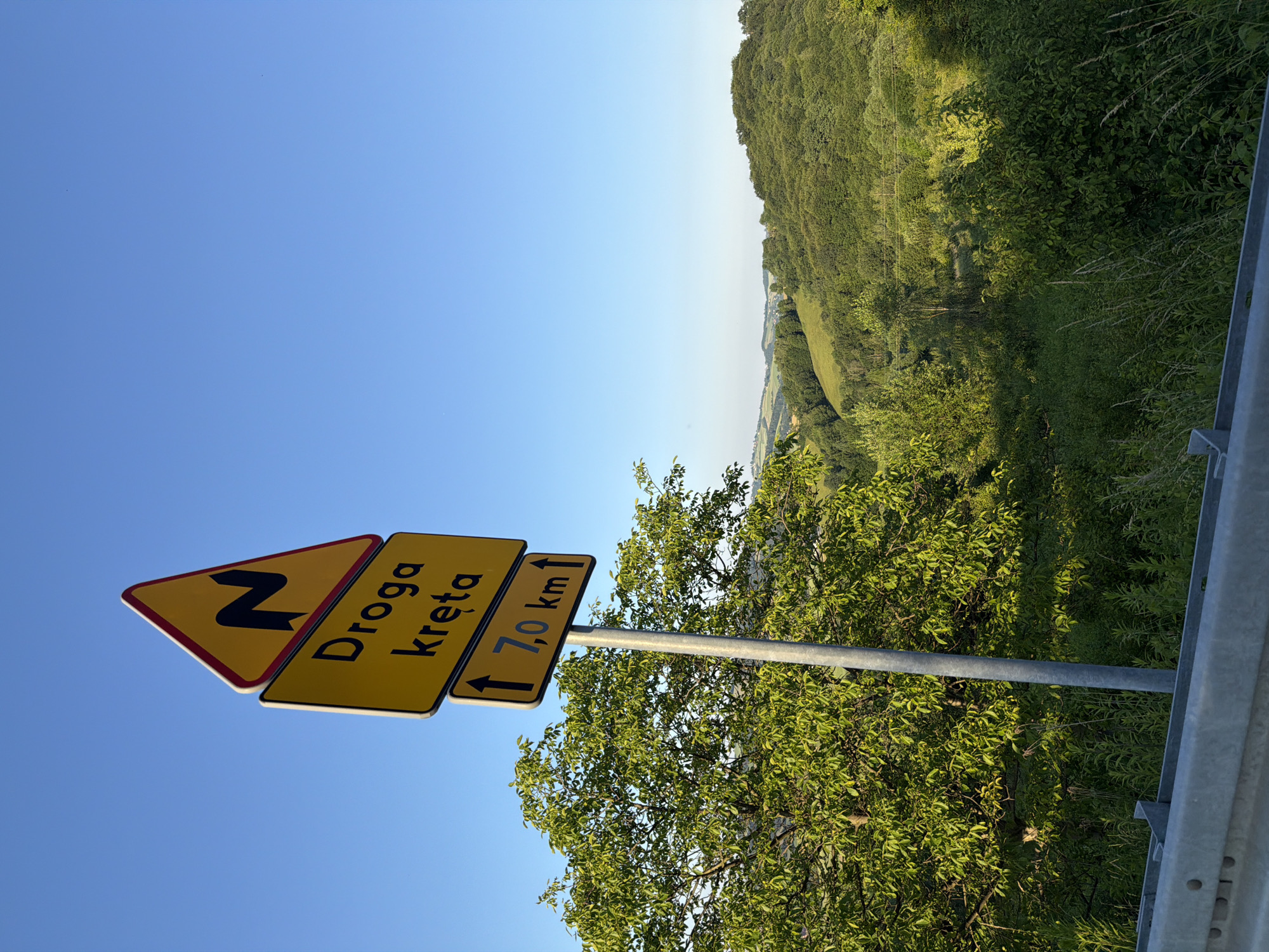 This is a medium-shot, outdoor photograph taken from a roadside perspective. The scene is dominated by a road signpost on the left, with three yellow signs stacked vertically. The uppermost sign is triangular with a red border and depicts a black, zig-zag symbol indicating a winding road ahead. Below this is a rectangular sign with black text that reads "Droga kręta," which translates to "Winding road." The bottommost rectangular sign displays two black arrows pointing upwards, flanking the text "7,0 km," indicating the distance to the end of the winding road.
The road signpost is a metallic grey pole, and to its left, a lush green tree with dense foliage is prominent. The background reveals a rolling, hilly landscape characterized by green fields and dense forests under a clear, bright blue sky. The overall impression is a rural or semi-rural setting, likely in Poland given the Polish text on the sign. No people or animals are visible. The lighting suggests it is daytime, with sunlight casting shadows and illuminating the landscape. A silver metal barrier is visible in the foreground, suggesting the photograph was taken from alongside a road or overlook.