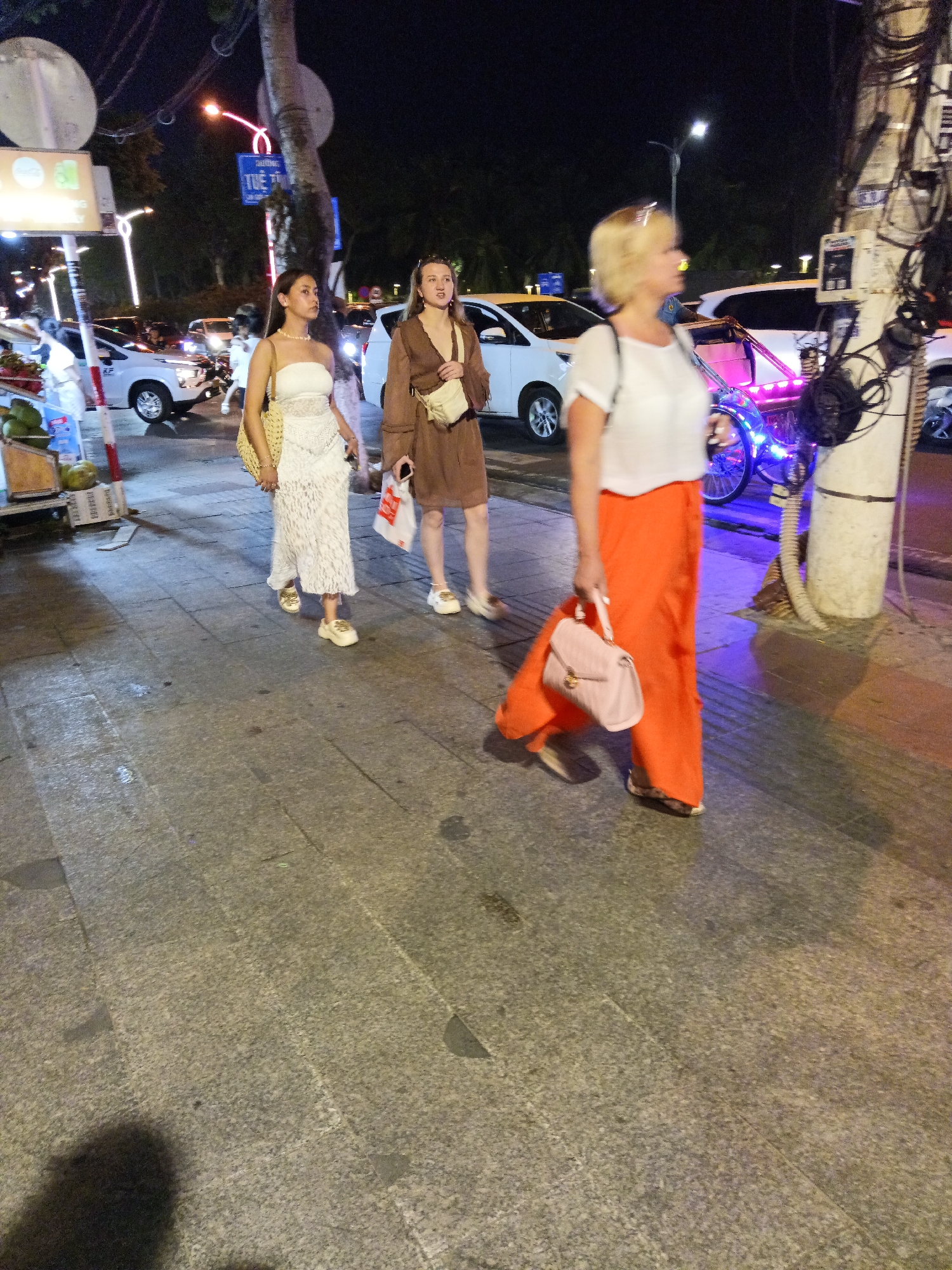 The image depicts a nighttime scene on a paved sidewalk in Nha Trang, Vietnam. Three women are walking. The woman on the left wears a white strapless dress and carries a straw bag. The woman in the middle wears a brown dress and a light-colored crossbody bag, holding a white paper bag in her right hand. The woman on the right has blonde hair and wears a white top and a flowing orange skirt. She carries a pink handbag.
In the background, several cars are visible, including a white car with its headlights on. A bright pink and blue light emanates from what appears to be a decorative rickshaw or similar vehicle. A utility pole with numerous wires and a meter is prominent on the right side of the frame. Streetlights illuminate the scene. The ground is paved with large, grey tiles.
The time of day is night, and the lighting suggests a warm, humid climate. The overall mood is casual and urban, with the women appearing to be enjoying an evening stroll.
Visible text includes "TUE 10" on a blue street sign in the background. Other text on signs and vehicles is not clearly legible.