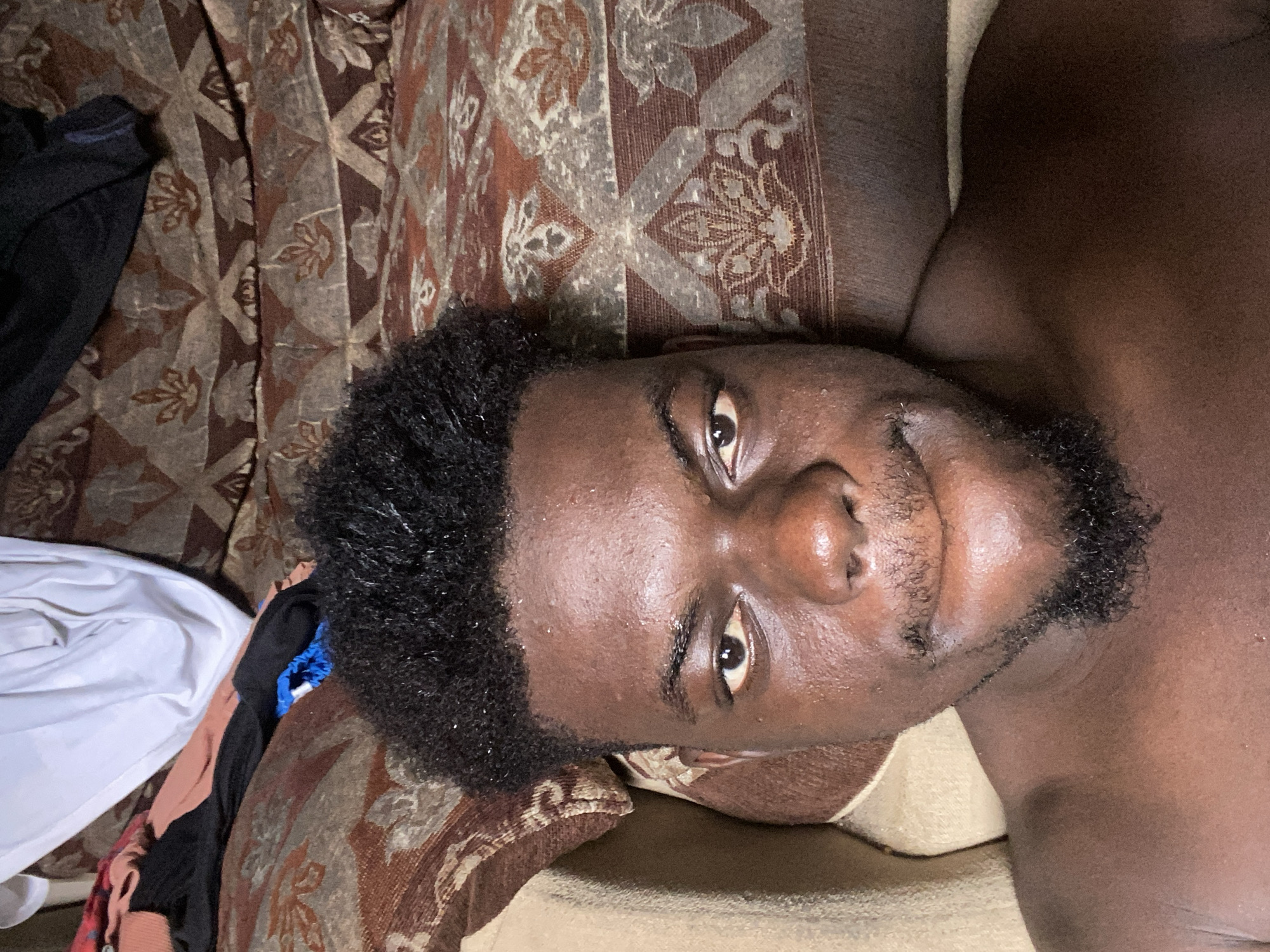 A dark-skinned male lies on a patterned brown and beige fabric surface. He has short, dark, curly hair and a short, dark beard. The background includes clothing items in various colors, including white, black, and pink. The location appears to be indoors, with the man lying on a bed or sofa. The city location is unknown.