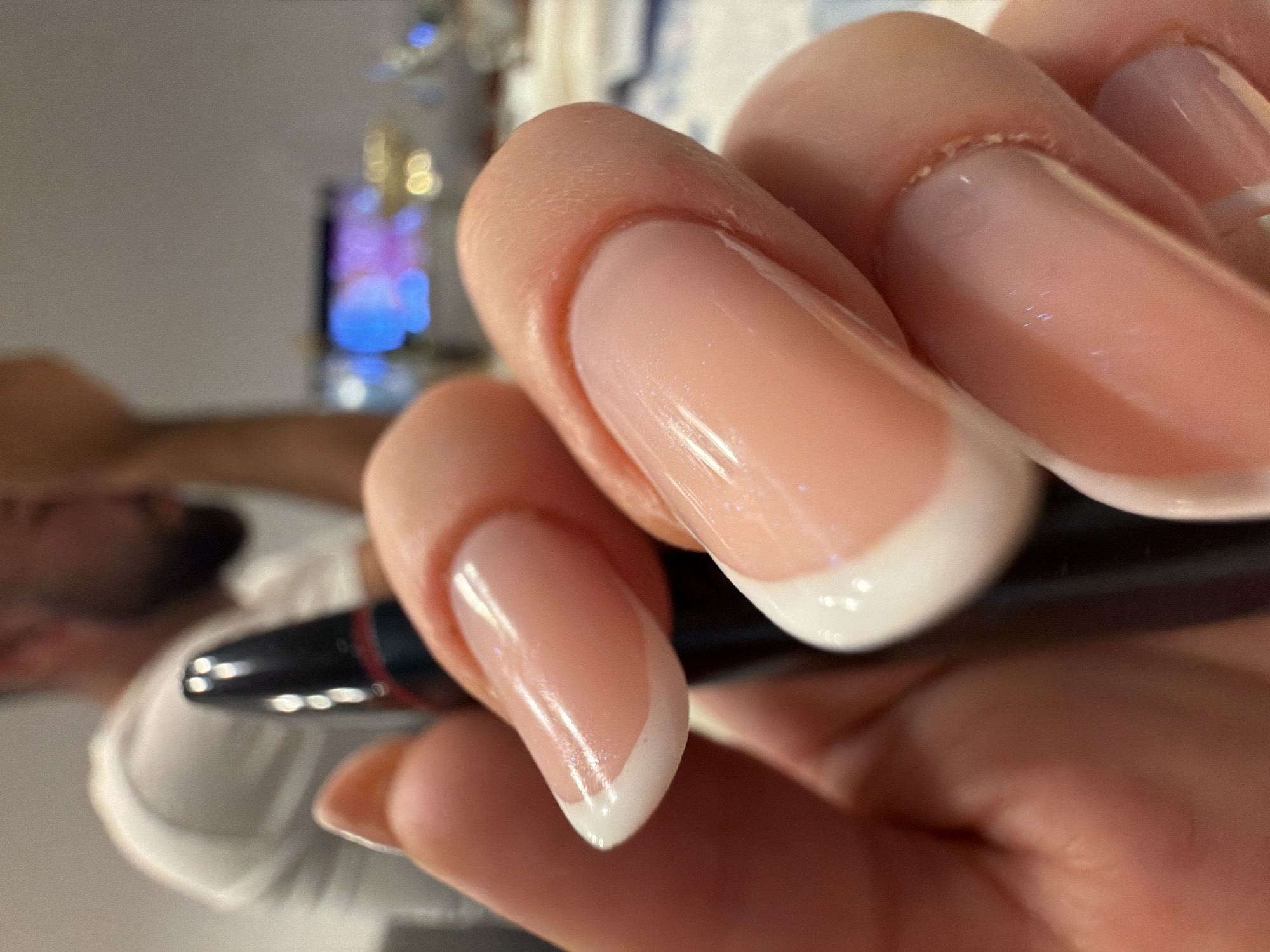 In a close-up, low-angle shot, a person's manicured hand, adorned with pink acrylic nails featuring a classic French tip, grips a black pen with a red accent. The pen is positioned vertically, with its tip pointing upwards and slightly to the left. The background is blurred, showing a hint of a person in a white t-shirt, out of focus, and a colorful, out-of-focus display in the distance, possibly a screen. The overall setting appears to be indoors, in what could be a casual environment. The image does not contain any information that could be used to identify its location in Florești, Romania.