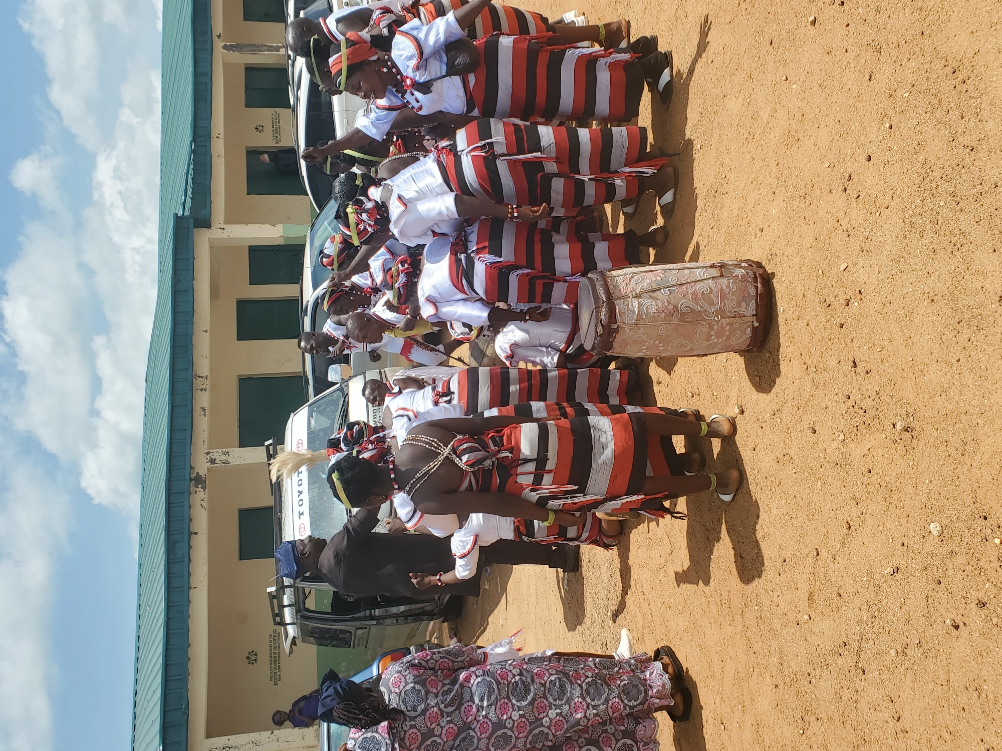 In the city of Kabong, Nigeria, an outdoor gathering is underway in front of a light-colored, two-story building with a green roof. The building appears to be a public facility or school, with multiple closed doors and windows. In the foreground, a group of people dressed in traditional attire is congregated on a sandy surface. The attire features black, white, and red striped skirts or wrappers, paired with white tops. Some individuals have beaded necklaces and headbands. The group appears to be in motion, with some individuals dancing or raising their arms. A man in a dark suit and a blue cap stands near a white van, possibly a Toyota, with a luggage rack on top. A drum, decorated with a floral pattern, is visible in the lower left of the frame. The sky above is partly cloudy, suggesting it is daytime. The overall atmosphere suggests a cultural event or celebration.