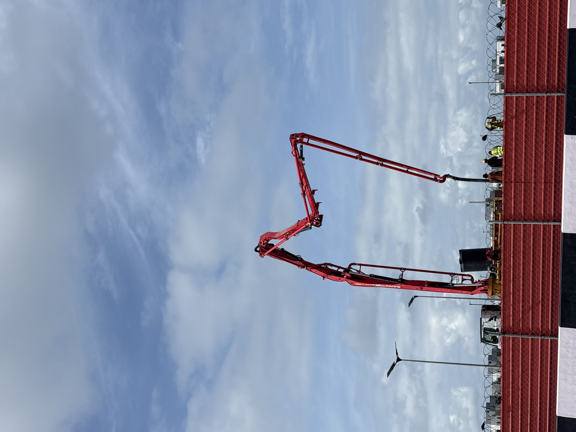 This image captures a daytime construction scene in Igboefon, Nigeria, dominated by a large, red concrete boom pump set against a partly cloudy sky. The photograph is taken from a slightly low angle, looking towards the construction activity.
The central and most prominent object is a bright red concrete pump truck, identifiable by the "SANY" logo visible on its articulating arm. Its multi-jointed boom is fully extended upwards and towards the left side of the frame, poised for or engaged in concrete pouring.
In the mid-ground, a tall red corrugated metal fence topped with several rows of barbed wire runs horizontally across the right half of the image. A black and white striped barrier or post is positioned at the far right edge of the fence. Behind this fence, several construction workers are visible. They are wearing standard personal protective equipment, including high-visibility vests (some yellow, some green) and hard hats, suggesting they are actively involved in supervising or performing tasks related to the concrete operation. A black tank, possibly for water or fuel, and the cab of another construction vehicle (likely a concrete mixer or support truck) are also partially visible behind the fence.
In the lower-left quadrant, a modern street light or utility pole with a dark, flat head stands against the sky. The sky itself is a bright blue, interspersed with scattered white cumulus and cirrus clouds, indicating fair weather conditions suitable for outdoor work. The overall impression is one of active industrial construction taking place under clear daylight.