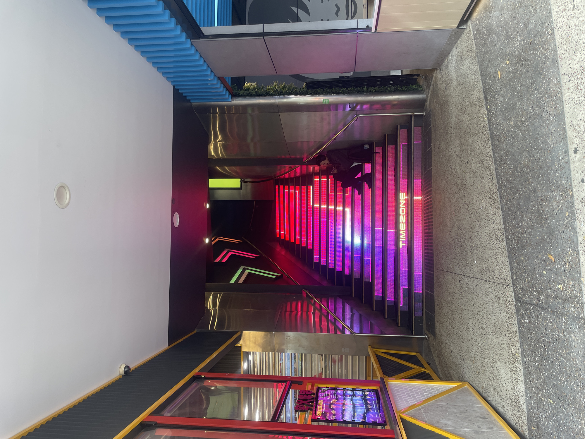 In Surfers Paradise, Australia, an exterior view shows a bright, modern entrance to a venue. The main focal point is a wide staircase illuminated with vibrant pink and purple LED lights on each step. The stairs are bordered by sleek, metallic walls that reflect the colorful light. On the left side of the image, a section of a building facade features a red and black exterior with a yellow trim. A digital display board, showing images of people and text, is visible. A security camera is mounted on the dark ceiling above this entrance. A lone individual, dressed in dark clothing, is seated on the illuminated staircase, looking down and holding a phone. Above the staircase, neon lights in the shape of arrows, in green and pink, are mounted on the dark ceiling. To the right of the staircase, a portion of a building facade with blue vertical slats is visible, and a large, stylized black and white graphic of a face is partially seen on the wall. The ground in front of the entrance is paved with speckled concrete.