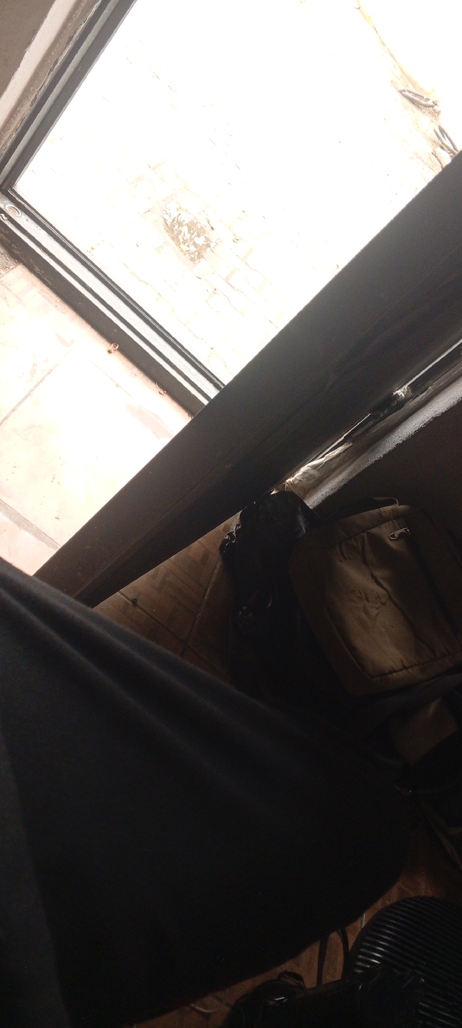 This image captures a close-up view of a doorway or window frame, looking outwards towards a bright, white, possibly overcast sky. The frame itself appears to be dark metal or wood. Below the frame, on the interior side, several objects are visible.
To the left, a large expanse of dark fabric, possibly clothing or a blanket, dominates the foreground. To the right of the fabric, a tan-colored bag with a flap and buckle is positioned on what appears to be a wooden surface. In front of the bag, a dark, unidentifiable object, possibly a small animal or a pile of dark material, is situated on the same surface. The surface itself appears to be made of wooden planks or a similar material.
The scene is dimly lit, with the brightest area being the sky visible through the opening. There is no clear indication of the specific location within Ilado, Nigeria, or any definitive action taking place. The image is a still photograph with no discernible movement or interaction between subjects.