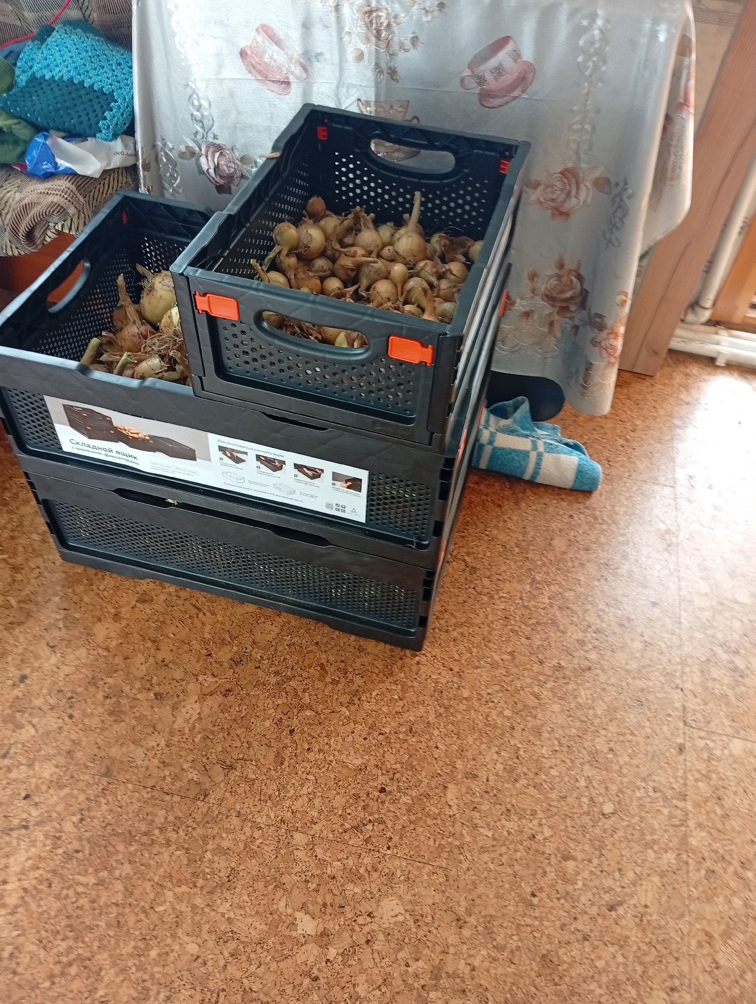 The image shows two stacked black plastic crates filled with harvested onions. The crates are placed on a cork floor, and a tablecloth with a floral pattern hangs in the background. A blue and white striped towel is folded on the floor to the right of the crates. The scene appears to be indoors, possibly a home.