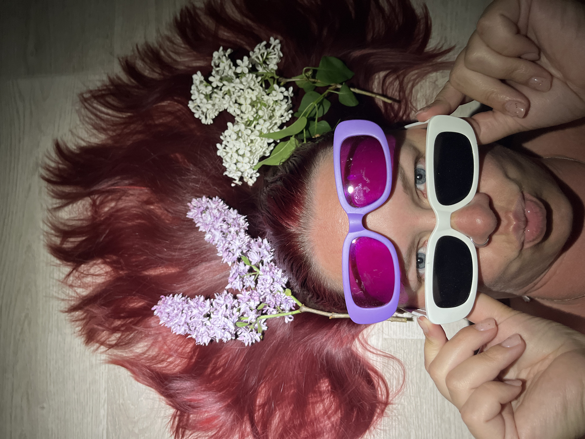 The image depicts a person lying on a light-colored wooden floor. The person has long, reddish-brown hair spread out around their head, adorned with sprigs of white and purple lilac flowers. The person is holding two different pairs of sunglasses in front of their eyes: the left pair has purple frames and magenta lenses, while the right pair has white frames and black lenses. The person is sticking their tongue out slightly. The location is Chaikovsky, Russia.
