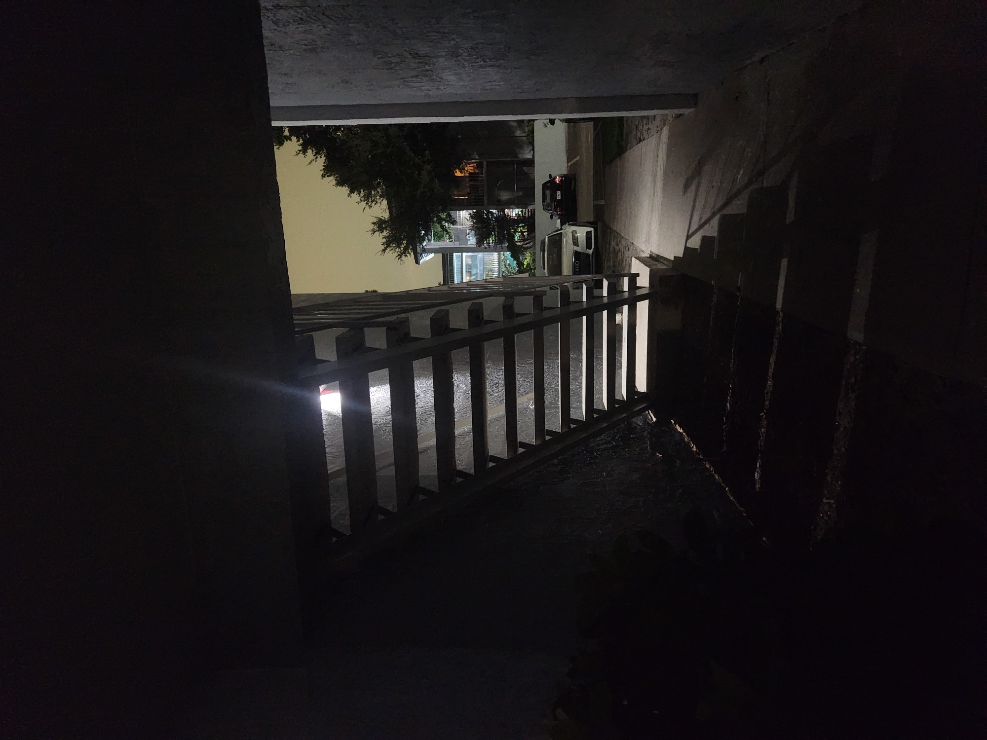 The image depicts an exterior stairway at night. The stairway is constructed with metal treads and risers and a metal handrail. The lower portion of the stairway is in deep shadow, with some steps illuminated by ambient light. Above the stairway, a bright light source is visible, possibly an exterior light fixture. To the right of the stairway, a concrete walkway leads to a parking area. In the parking area, two cars are visible: a white sedan and a dark-colored sedan. In the background, apartment buildings and trees are dimly lit by the night sky, which appears overcast and yellowish. The overall scene suggests an apartment complex in Altamonte Springs, United States of America. There are no people visible in the image.