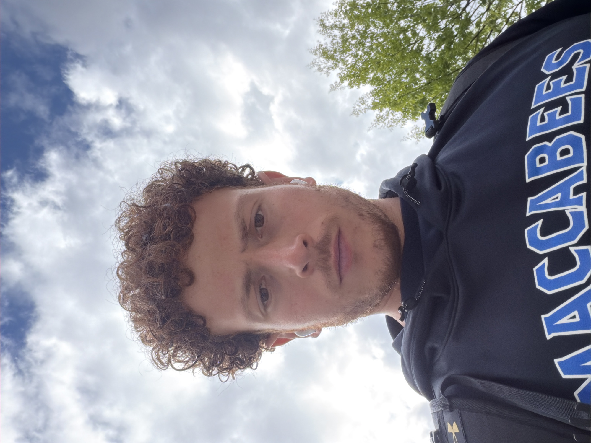 A selfie-style, angled upward shot shows a fair-skinned man with curly brown hair, a short beard, and wearing an earbud in his left ear. He is wearing a dark blue hooded sweatshirt with the word "MACCABEES" printed in large, blocky white letters with blue outlining down the right side of the image. A black Under Armour bag strap is visible on his right side. Above him is a view of a cloudy sky with patches of blue and a few green tree branches peeking into the top right corner. The image doesn't provide sufficient information to confirm the location in New York City.