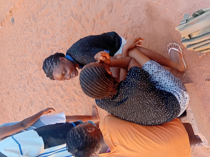 Three girls are resting on a sandy surface. One girl, wearing a black patterned shirt and checkered shorts, sits behind another girl in an orange shirt. A third girl, wearing a black and white striped shirt, rests on the sand with one arm outstretched. One girl wears a black shirt with blue trim and has braided hair. Sand and bags are visible.