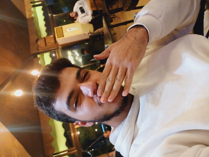 A person with short, dark hair and a trimmed beard is facing the camera, partially covering their mouth with their hand. The individual is wearing a white hooded sweatshirt. The scene appears to be indoors, possibly a restaurant, with soft lighting. In the background, other people can be seen, along with window reflections and restaurant decor. The image is slightly tilted, adding a casual feel.