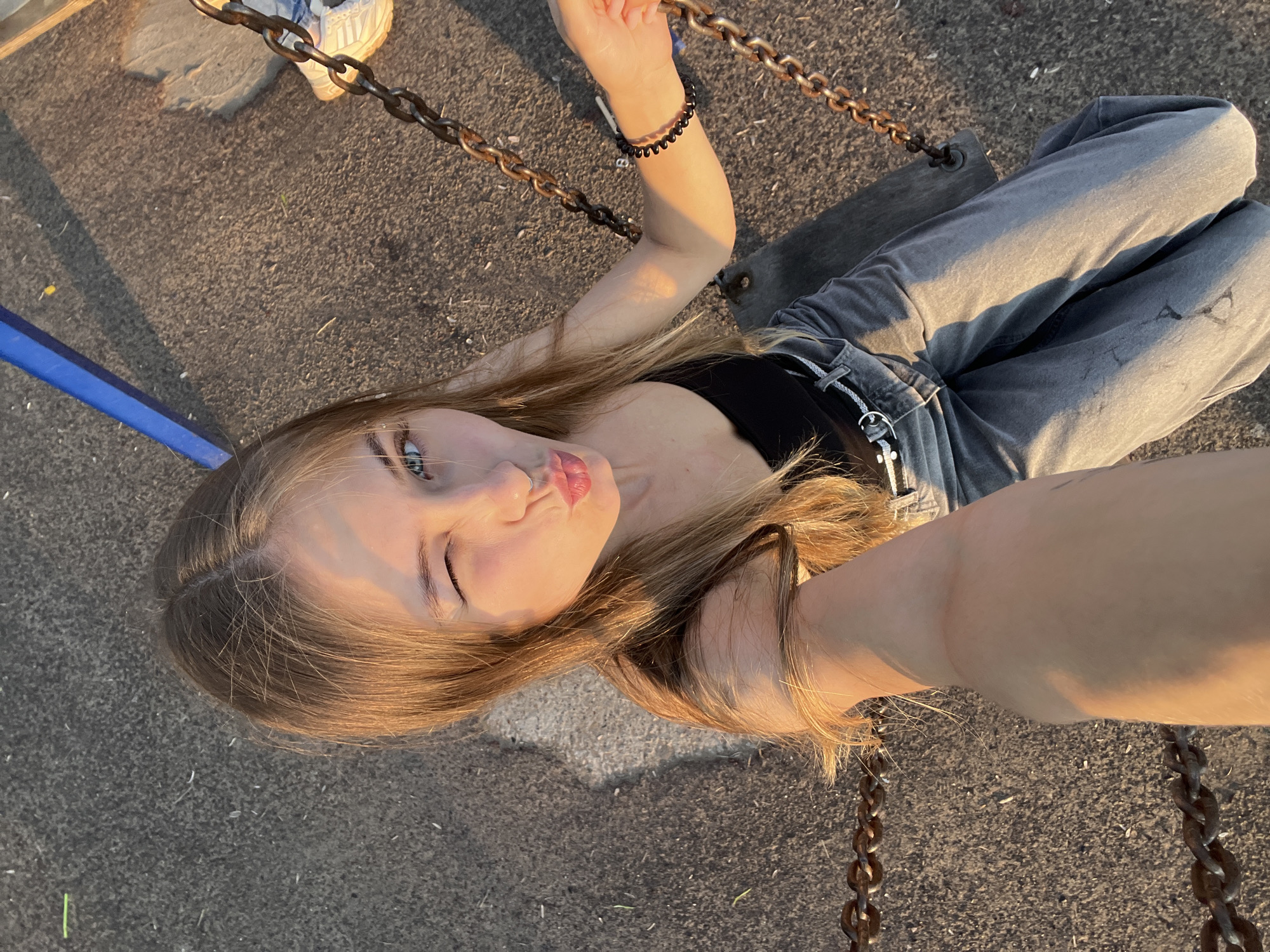 This media file features a young woman in a park setting, possibly during the golden hour due to the warm, directional lighting. She is sitting on a swing, evident from the chains and wooden seat visible in the frame. The ground appears to be a mix of asphalt and gravel, typical of outdoor recreational areas.
The woman has long, blonde hair and is wearing a black tank top and light-wash jeans. She is looking directly at the camera, with one eye winking and her lips puckered in a playful expression, suggesting a lighthearted and possibly fun mood. She is wearing a black beaded bracelet on her left wrist. Her right arm is visible, resting on the swing chain. In the background, a blue support beam of the swing set is partially visible, along with a white sneaker on her left foot.
The scene is outdoors, with the sunlight casting long shadows and highlighting the texture of the ground and the woman's skin and hair. There is no visible text in the image. The overall impression is one of youthful energy and enjoyment of a casual moment outdoors.