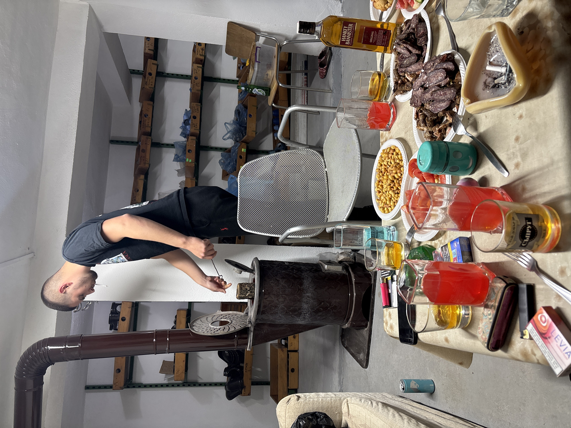 An adult male stands to the left of a wood-burning stove with a stovepipe, apparently stoking it, in a room that appears to be located in Zetiovo, Bulgaria. The room has shelving on the walls filled with various items. A table laden with food and drinks, including a bottle of "Hankey Bannister" whiskey, several glasses of beverages, plates of meat and nuts, and an ashtray filled with cigarette butts, sits in the center of the room. A metal chair is tucked under the table. A portion of a light-colored sofa is visible in the foreground.