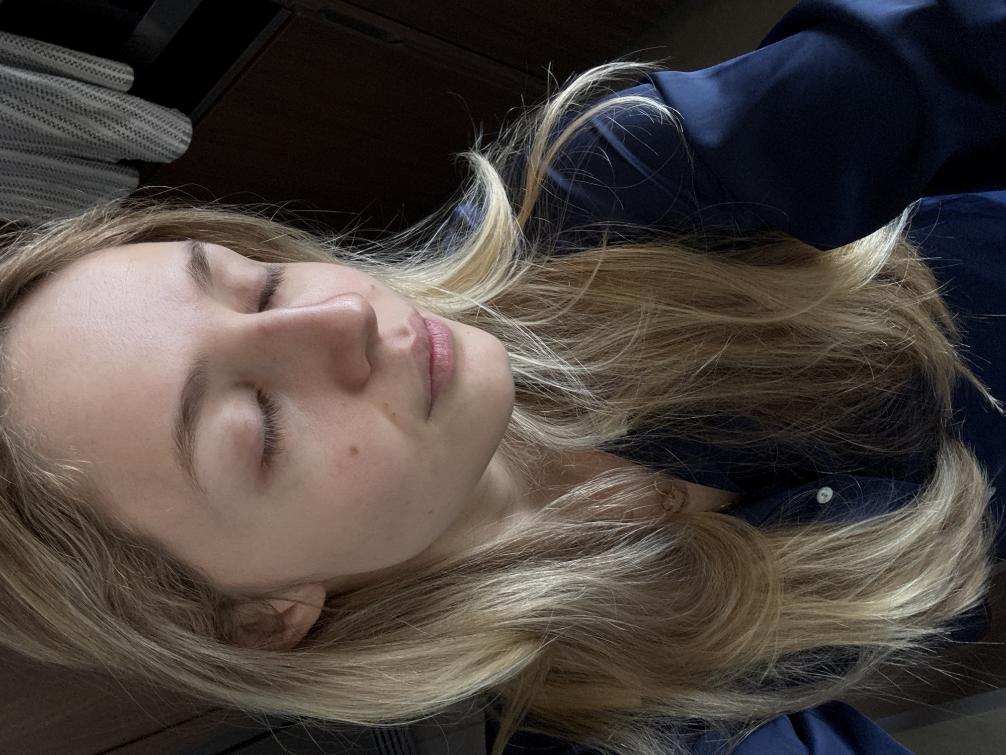 A blonde-haired woman lies with her eyes closed, possibly sleeping. She wears a dark blue garment, perhaps a blouse or jacket. Her head rests on a dark surface. The environment includes what appears to be a wooden cabinet or dresser behind her, with a glimpse of a gray and white patterned fabric or item on top. The photo does not contain any information that would allow the specification of a location in Mexico City, Mexico.