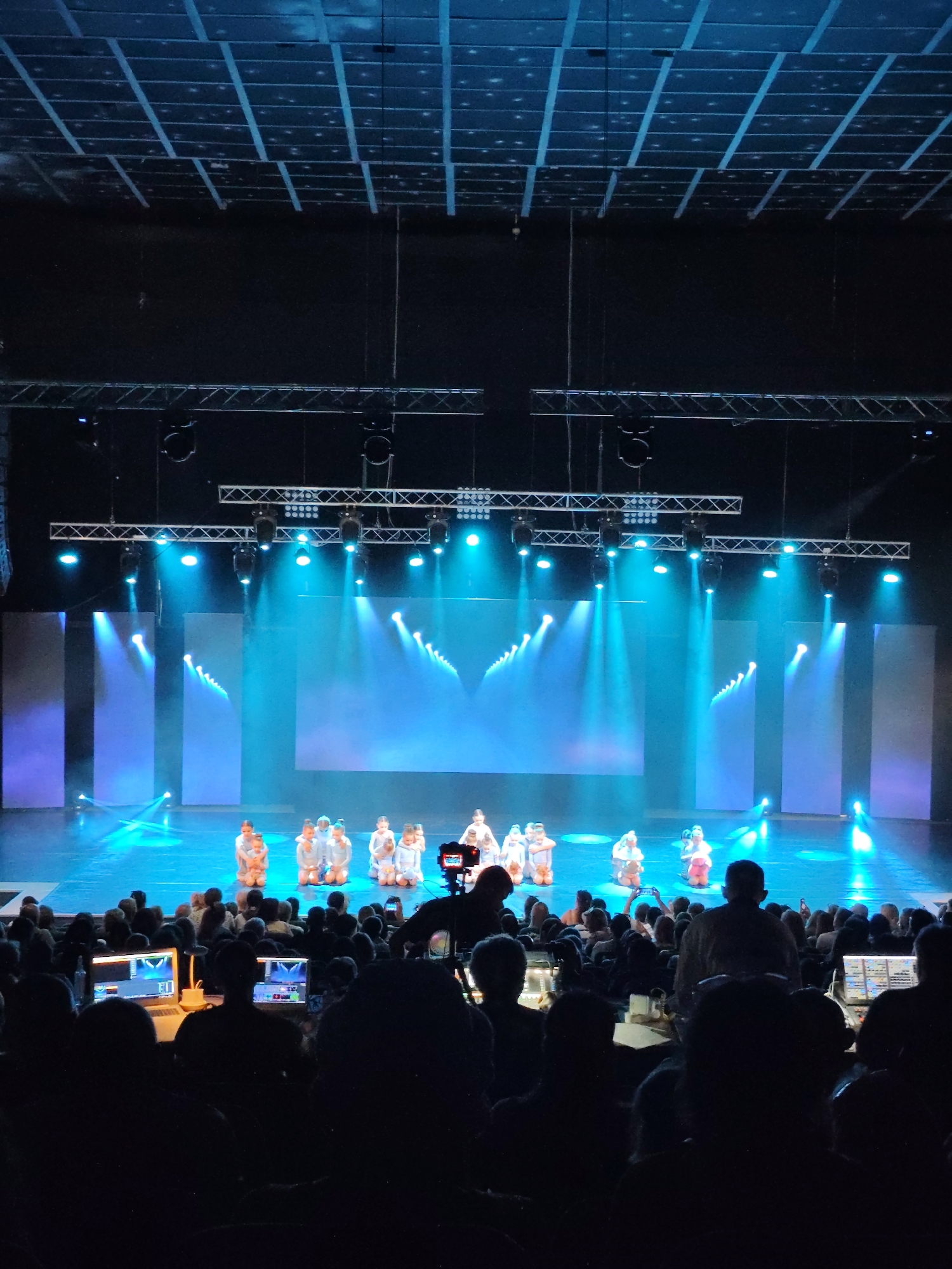This image depicts a live performance on a stage with a large audience in the foreground. The stage is illuminated with blue and white lights, creating a dramatic effect. Several children, dressed in light-colored costumes, are positioned on the stage, some kneeling and others standing. The background of the stage features large screens displaying abstract patterns of light. Above the stage, a complex rigging system with lights and sound equipment is visible.
In the audience area, many people are seated in the dark, with some individuals in the front rows engaged with laptops and other electronic devices, suggesting they might be operating technical equipment for the performance. The overall atmosphere suggests an indoor event, possibly a dance recital or a theater production.
The description provided does not include information about the specific location in the city of Mykolaiv, Ukraine, as that detail is not present in the image itself.