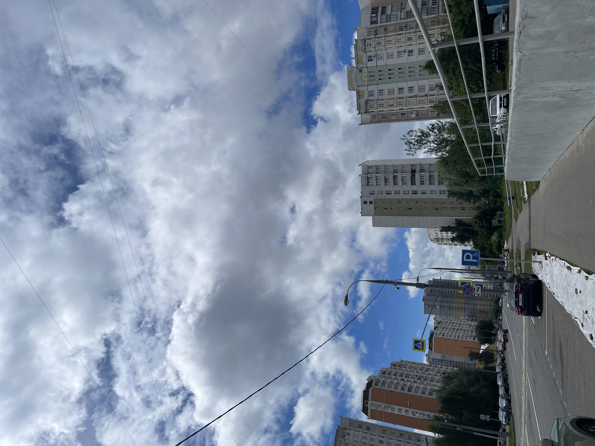 This is a street-level, eye-level, medium shot photo of a city street in Moscow, Russia. The image features a modern, multistory apartment buildings with balconies on the left and right sides of the frame. The sky is partially cloudy with blue patches, and power lines are visible strung across the top of the frame. A street lamp is present on the left side of the road. Traffic signs, including a pedestrian crossing sign, a "no parking" sign, and a disabled parking sign, are visible on the right side of the street. Several cars are parked along the side of the road, and some are driving on the road. A sidewalk runs along the right side of the street, with a low concrete barrier and a metal railing. The overall scene depicts an urban residential area on a cloudy day.