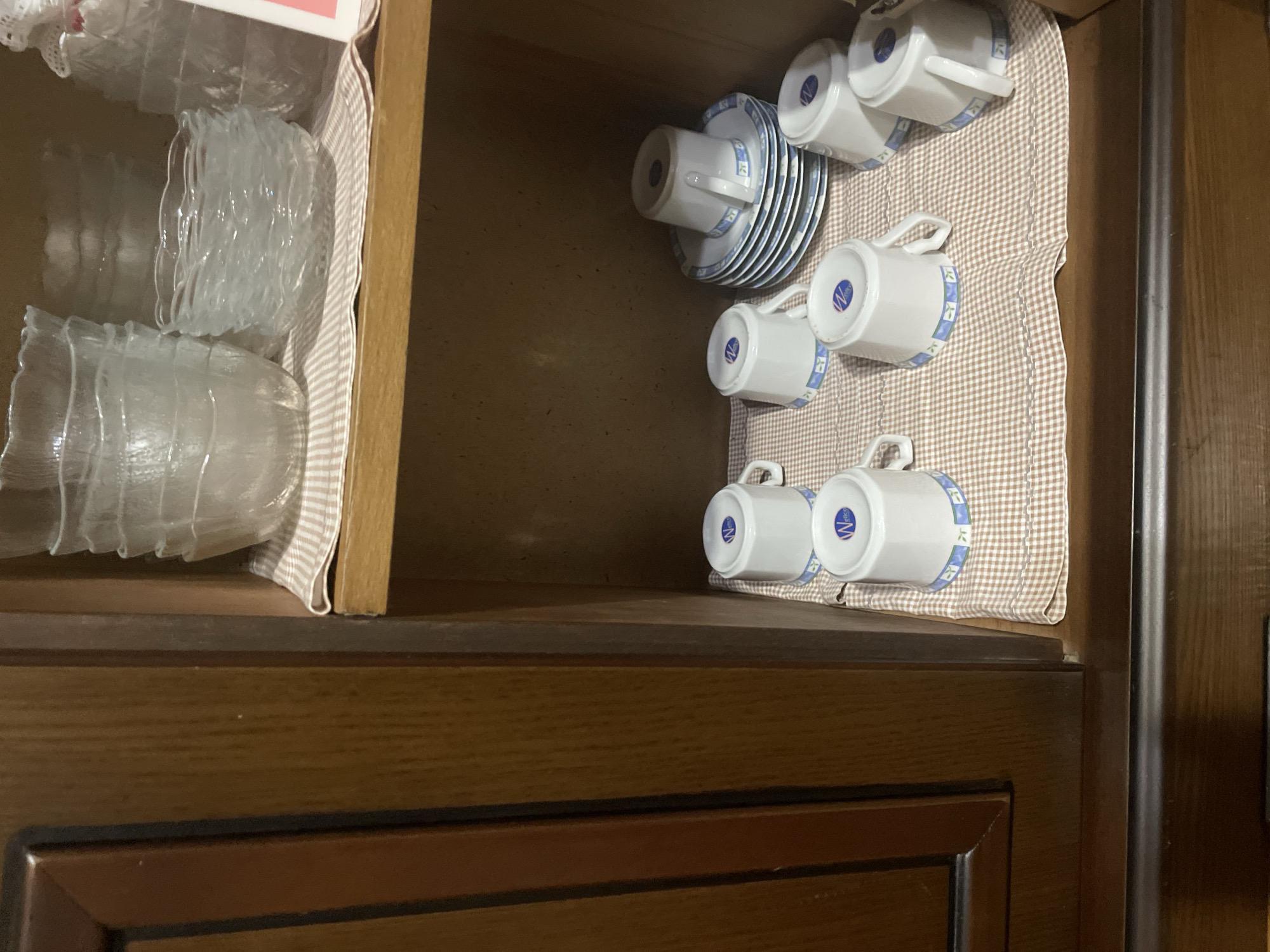 The image shows the interior of a wooden cabinet. The top shelf contains stacked clear glass bowls and a stack of clear glass dishes. The lower shelf contains several white ceramic coffee cups with a blue and green decorative band around the rim, and a stack of matching saucers. The cabinet appears to be in a home or retail setting. There is no indication of its specific location in Kadıköy, Turkey. The scene is static and no people or actions are depicted.