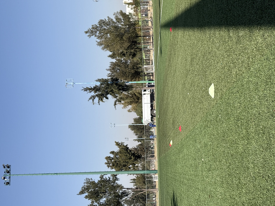 The scene depicts an artificial turf sports field located in a sports complex in León, Mexico, under a clear blue sky. In the foreground, the green artificial turf is marked by four disc cones: one light green and three red. A long, dark shadow extends from the lower right corner towards the center of the field. In the midground, a chain-link fence separates the playing surface from an asphalt or paved service area.
A white service truck, appearing to be a tanker or utility vehicle, is parked parallel to the fence on the paved area. Two individuals are present near the truck; one, dressed in a blue uniform, stands to the left side of the truck, while another individual, also in work attire, is partially visible to the right of the truck. Their actions suggest attendance to the vehicle or the immediate surroundings.
Beyond the fence and truck, multiple tall poles rise against the sky. On the far left, a prominent green pole equipped with several floodlights at its apex is visible. To the right, another tall, slender pole, painted blue and topped with a square framework, stands amongst a line of dense green trees that form the background. Additional blue-painted light poles are discernible in the distance, further integrating the scene into a structured outdoor recreational facility. A light-colored building is faintly visible in the far right background, behind the trees.