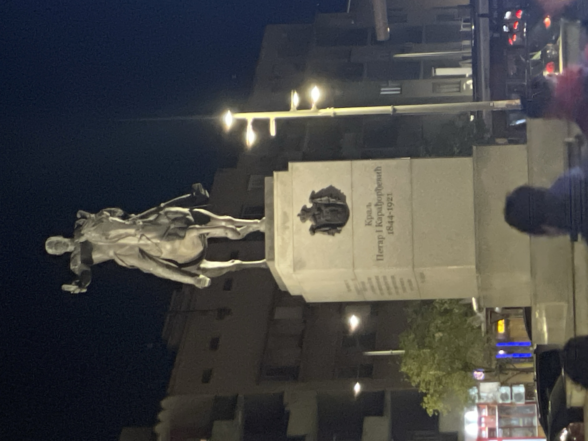 The image depicts the monument to King Peter I Karadjordjevic in Novi Sad, Serbia, at night. A bronze equestrian statue stands atop a white stone pedestal, with the inscription "Краљ Петар I Карађорђевић 1844-1921" (King Peter I Karadjordjevic 1844-1921). The monument is illuminated by streetlights. Buildings are visible in the background, as well as what appears to be a commercial area with stores and people. Some people are standing close to the monument, and others are passing by.