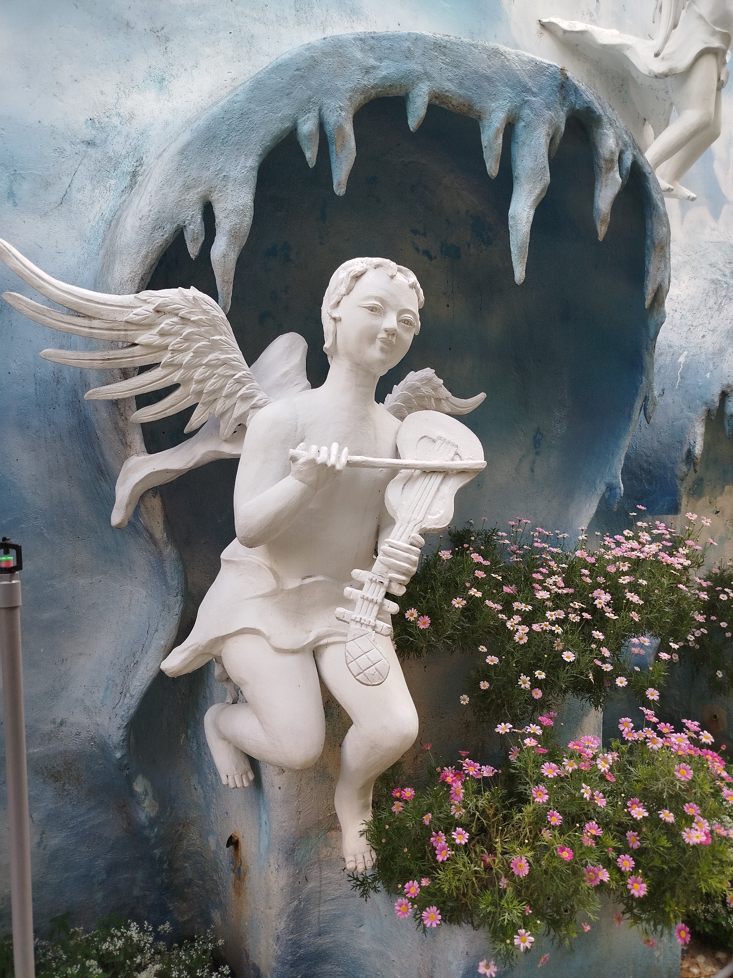 A statue of a winged angel playing a lute is partially embedded in a blue and white grotto with stalactites, which is surrounded by pink and white daisies. The statue is white. A portion of another statue is visible in the upper right corner. The foreground includes a gray pole. The photo was taken in Da Lat, Vietnam.