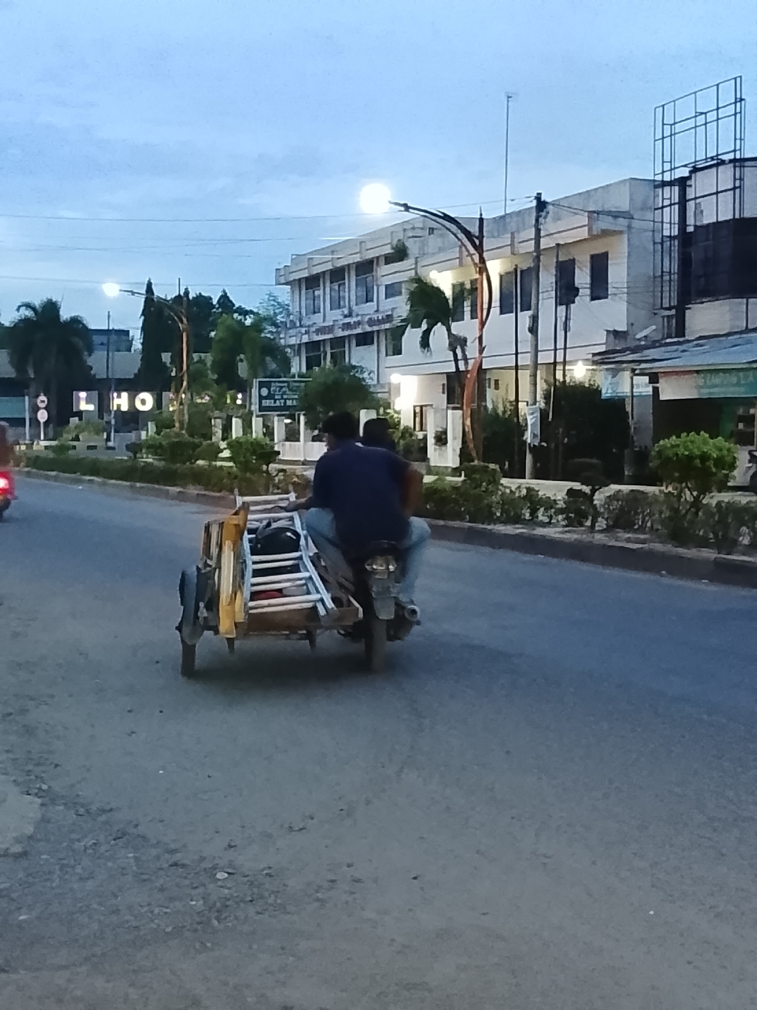 In Lhokseumawe, Indonesia, on a paved road with a grey hue and scattered small stones, two individuals are traveling on a motorcycle. The motorcycle is towing a small, yellow trailer carrying a ladder and some other items. The passenger is sitting in the trailer, while the driver is on the motorcycle seat. They are moving away from the camera. In the background, a multi-story building with a white facade and dark windows is visible, along with some trees and streetlights. The sky is overcast with a pale blue color. The lighting suggests it is either early morning or late evening.