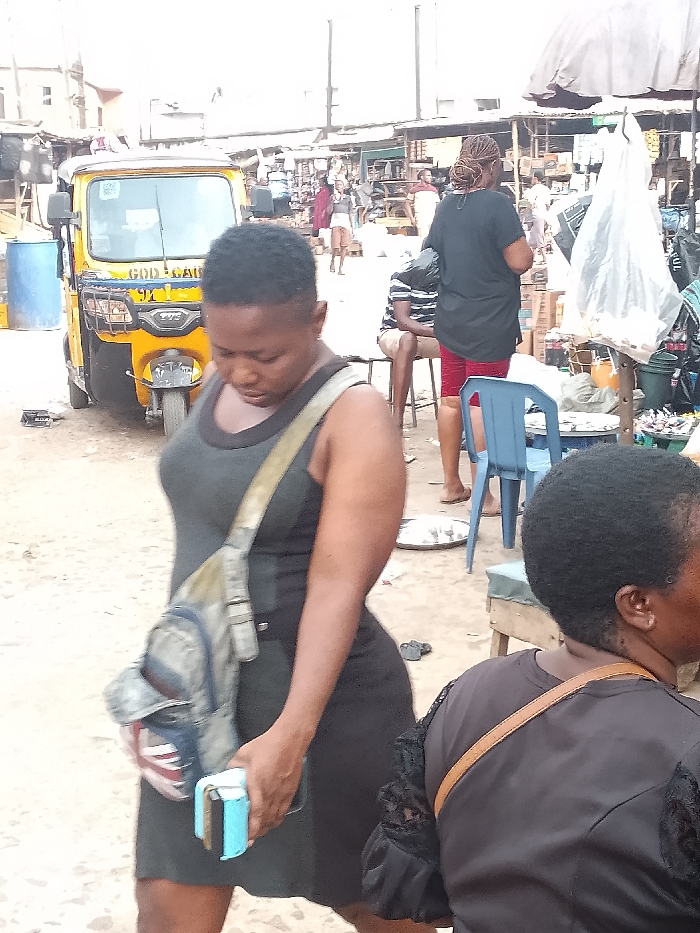 The image captures a vibrant and busy market scene in what appears to be Akpaka Forest Reser, Nigeria. In the immediate foreground, a woman walks towards the left, her gaze directed downwards. She wears a dark grey or black sleeveless dress and carries a light-colored shoulder bag featuring a Union Jack design on one pocket. In her right hand, she holds a light blue object, possibly a wallet or phone, along with another dark item. To her right, the back of another person's head and shoulder is visible, wearing a black top with lace detailing on the sleeve and a brown bag strap across their shoulder.
The midground features a yellow motorized rickshaw, commonly known as a Keke NAPEP, with the words "GOD CALL" and "TVS" visible on its front. Several other individuals populate the scene; one person in a black shirt and red shorts stands facing away, while another in a striped shirt sits on a stool. A blue plastic chair is placed on the ground nearby. The background reveals a bustling market environment with numerous stalls, temporary structures, and more people, suggesting active commerce. The ground is a mix of dirt and sand. The bright, diffused lighting indicates it is daytime, possibly under an overcast sky or bright sun, without casting harsh shadows. The overall impression is one of a functional and lively outdoor market.