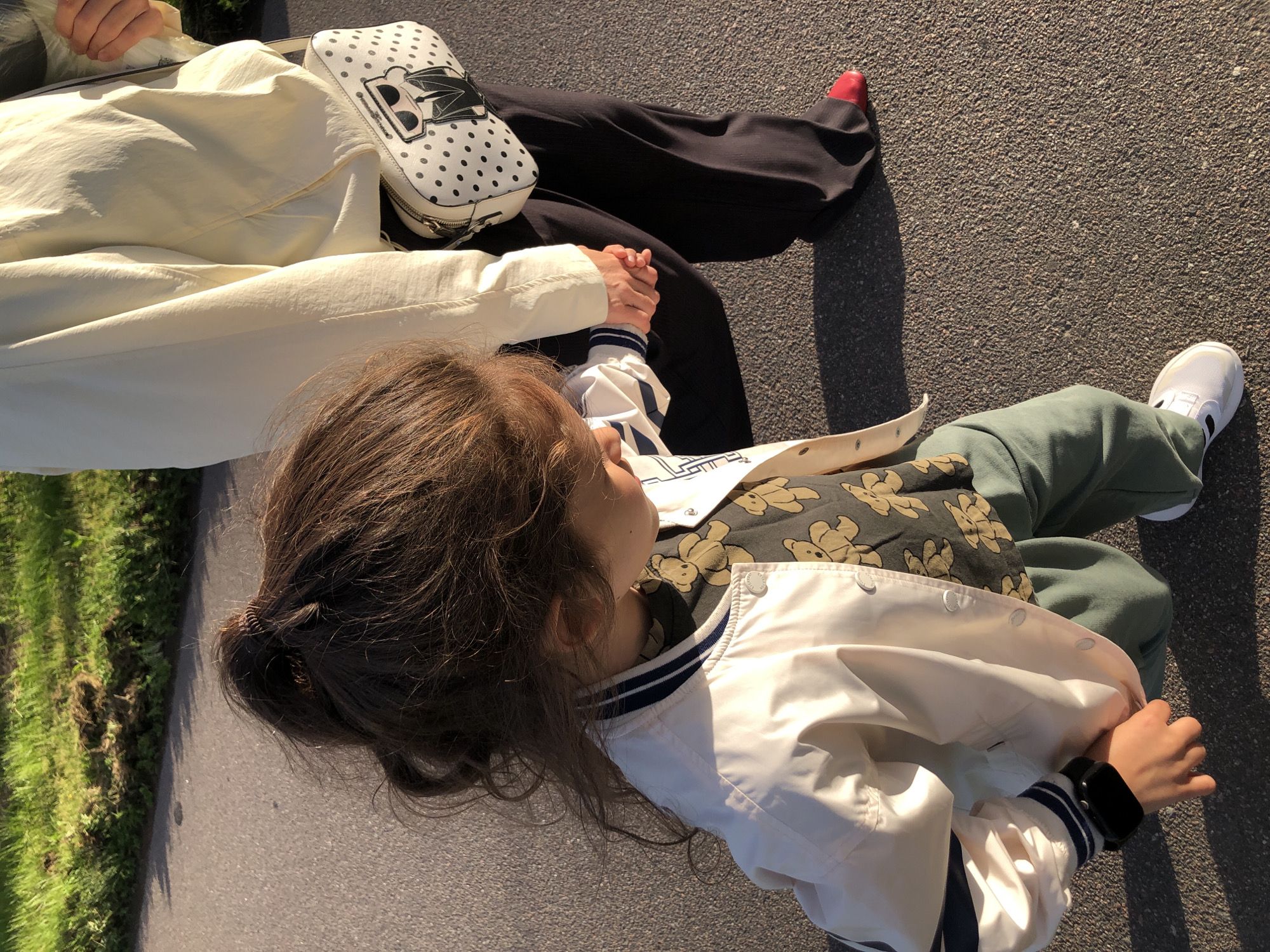 A child, seen from behind, walks hand-in-hand with an adult on an asphalt path. The child has long, dark brown hair tied in a ponytail and wears a white bomber jacket with blue and white striped cuffs and collar, over a dark green shirt with a teddy bear print, and light green pants. The child's left hand, wearing a black smartwatch, is clasped by the adult's right hand. The adult wears a light-colored jacket, dark pants, and red shoes. A black and white polka dot purse with a Karl Lagerfeld motif is slung over the adult's shoulder. The background shows a grassy area and a shadowed embankment. The image appears to be taken in Saint Petersburg, Russia, on a sunny day, likely in a park or along a waterfront promenade.