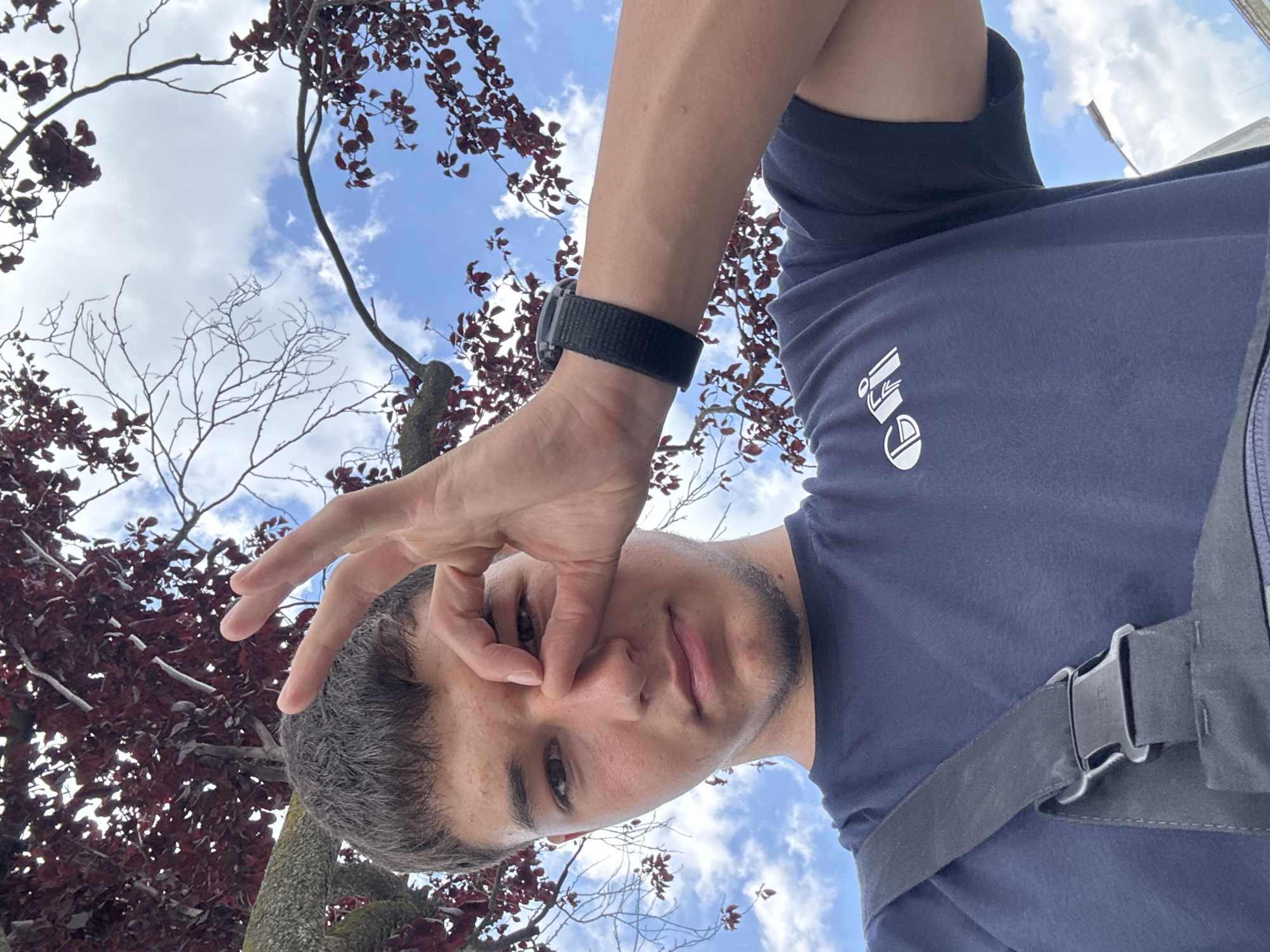The media file shows a close-up, low-angle shot of a young person looking up at the sky, with a hand held up to their face, obscuring part of it. The subject is wearing a dark blue t-shirt with a white logo on it and a black watch on their left wrist. The background is dominated by a blue sky with white clouds, and the branches of a tree with dark red leaves. The perspective suggests the photo was taken outdoors, possibly in a park or garden. The lighting indicates it is daytime, likely afternoon given the brightness. The overall mood is casual and playful. There is a strap with a buckle visible on the right side of the frame, which could be part of a bag or backpack. The logo on the t-shirt is difficult to discern clearly, but appears to be a stylized design. No other text is visible in the image. The location context is Mannheim, Germany.