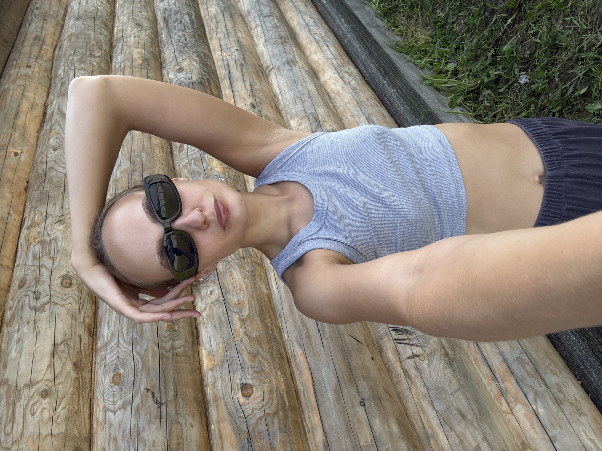 A young woman with blonde hair and sunglasses lies on a wooden surface. She is wearing a light blue tank top and dark shorts. Her left arm is bent at the elbow, and her hand is resting on her head. Her right arm is extended behind her. The wooden surface is made of logs placed side-by-side. To the right of the wooden surface is a patch of green grass and a dark, rough textured surface, possibly a fence or a border. The scene appears to be outdoors, as indicated by the grass and natural lighting. The woman has a relaxed pose, suggesting a moment of leisure. The time of day is likely daytime, given the bright lighting. The location is specified as Podshivalovo, Russia. There is no visible text in the image.