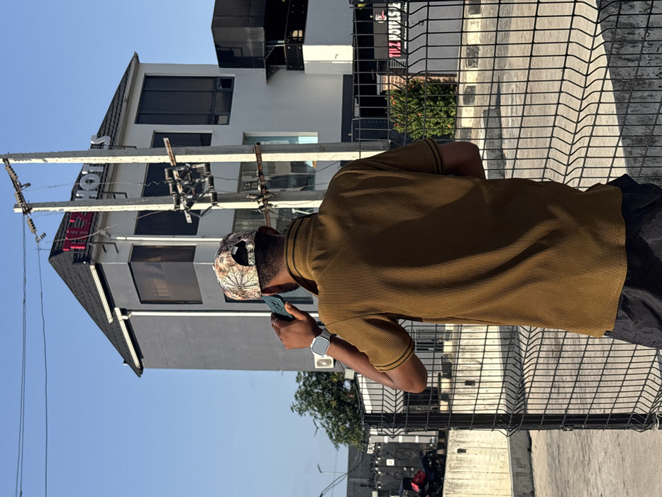 In Igboefon, Nigeria, a person is observed from behind, standing outdoors and holding a dark-colored smartphone to their right ear, appearing to be on a call. The individual wears a patterned baseball cap, a ribbed olive-brown short-sleeved collared shirt with dark trim on the sleeves, and dark shorts. A silver-toned smartwatch is visible on their left wrist. In the background, a multi-story building with grey walls, dark tinted windows, and a dark pitched roof is prominently featured. Red and white illuminated signage on the building's upper facade and roofline spells "TIZ HOTELS." A concrete utility pole, equipped with multiple horizontal cross arms, insulators, and electrical wiring, stands in the midground between the person and the hotel building. A black wire mesh fence occupies the immediate foreground. The ground surface is paved, and the sky overhead is clear and blue. Some green foliage is visible near the base of the hotel and behind the fence. A dark-colored motorcycle or scooter is partially visible parked behind the fence to the far left.