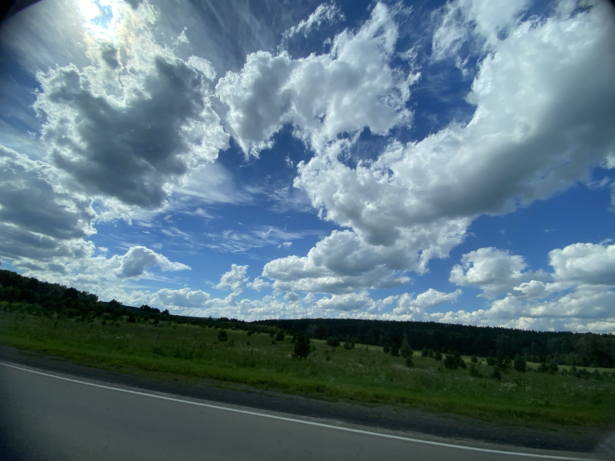 A wide-angle, low-angle shot captures a bright blue sky filled with dramatic, puffy white and gray clouds, some of which are illuminated by the sun, creating lens flare at the top left of the frame. The clouds are varied in size and density, with some appearing wispy and others thick and voluminous. Below the sky, a green, rolling landscape stretches horizontally across the frame. This landscape is covered in grass, with scattered trees and bushes, and a dense, dark green forest line forming the horizon. In the foreground, a portion of an asphalt road with a white line is visible on the left side, suggesting the viewpoint is from a vehicle traveling along it. The scene appears to be a rural or natural setting, possibly in the city of Tyulyugul, Russia, on a day with dynamic cloud cover. There are no people or animals visible in the image.