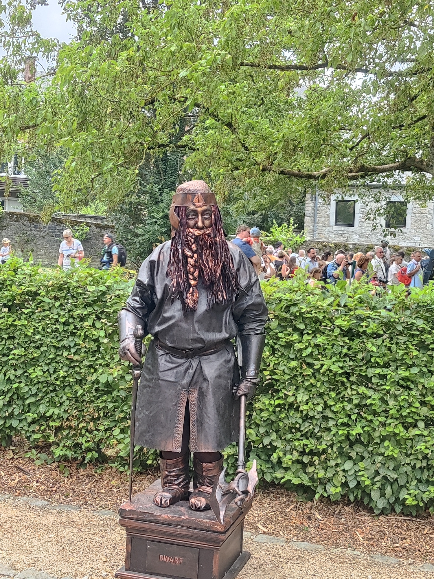 The photograph captures a living statue dressed as a dwarf, standing on a pedestal in what appears to be a public park or garden. The dwarf statue is positioned centrally in the frame, with a dense green hedge forming the background. Behind the hedge, a crowd of people are visible, suggesting an event or attraction. Further in the background, stone buildings and trees are present, indicating an outdoor urban or semi-urban setting. The dwarf is clad in dark, textured clothing resembling leather, with a helmet, a long braided beard, and holding an axe-like prop. The pedestal has the word "DWARF" written on it. The setting is outdoors, with dappled sunlight suggesting a partially cloudy day. The overall impression is that of a performance or display aimed at public entertainment in Marche-en-Famenne, Belgium, a city known for its medieval architecture and outdoor festivals.