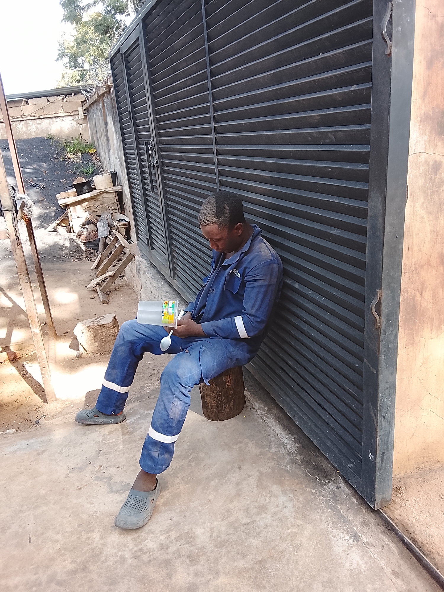A man in blue overalls with white reflective stripes on the legs sits on a wooden stump next to a large, dark metal gate. He is holding a clear plastic container with a white lid, which appears to contain a light-colored liquid. A spoon is also visible in his hand, and he is looking down at the container. His hair is short and appears to be dusted with a light-colored substance. The man is wearing grey slip-on shoes. The ground is concrete and dirt. In the background, there is a concrete wall and some building materials. The scene appears to be outdoors, possibly a workshop or industrial area, given the setting cues. The lighting suggests it is daytime, with shadows indicating the sun is present. The overall mood is one of a break or a moment of rest. No visible text can be discerned.