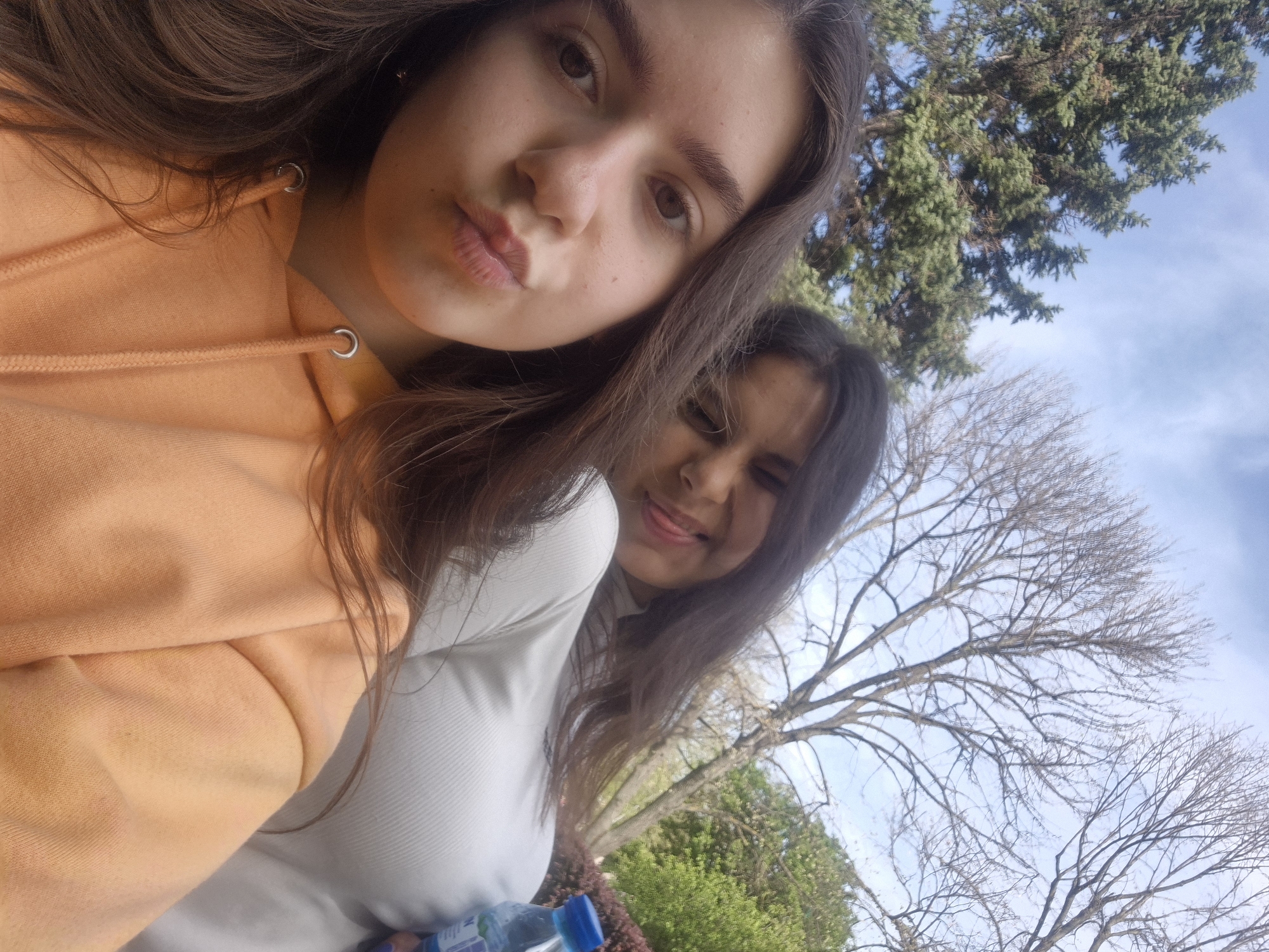 A tilted outdoor selfie features two young women in Pavlikeni, Bulgaria. The woman in the foreground has long, dark brown hair, light brown eyes, and is wearing an orange hooded sweatshirt. She looks directly at the camera with a slight pout. Behind her, a second woman with long, dark hair is partially visible, making a playful face by sticking out her tongue. She is wearing a white long-sleeved top.
The setting appears to be an outdoor park or natural area. The background consists of a clear blue sky with some faint clouds, visible between various trees. A large evergreen tree with dark green foliage is prominent in the upper background, while several bare deciduous trees, indicative of late autumn, winter, or early spring, are also present. A blue plastic bottle, likely containing water or another beverage, is partially visible at the bottom right, held by the second woman; text on its label is unreadable. The image captures a casual, friendly moment during the daytime.