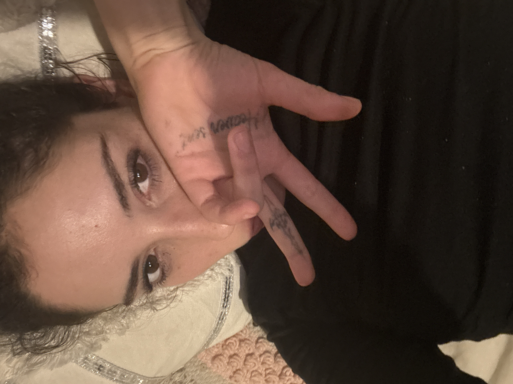 A woman lies on her side, seemingly on a pillow, with her dark hair spread out. She is wearing a dark-colored top. Her right hand is positioned in front of her face, with her fingers spread. Tattoos are visible on her hand and fingers: the words "Heaven sent" on the palm and a design on the finger. The background includes a white pillow with silver detailing and a textured pink surface. The location is not provided in the image.