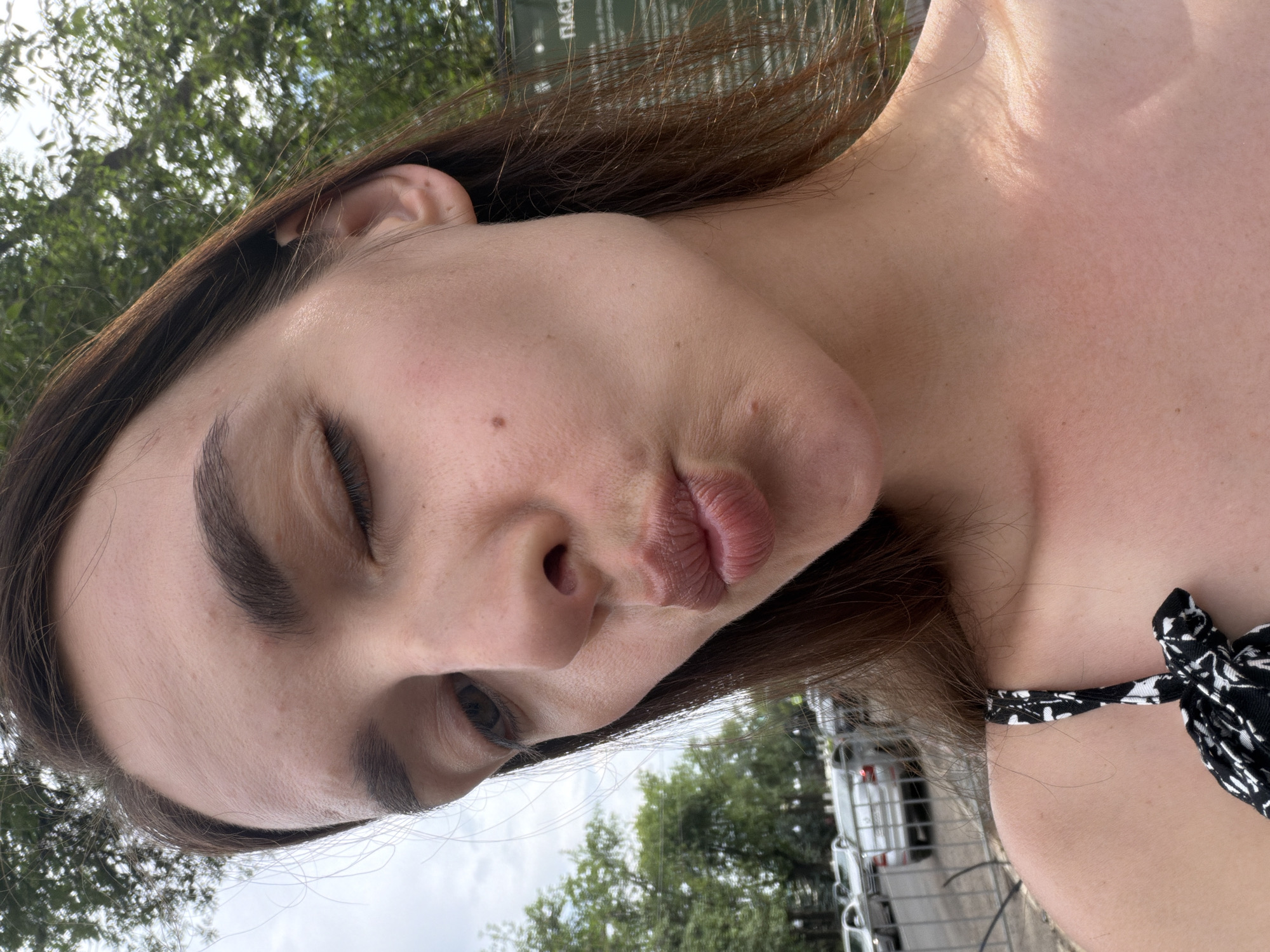 A young woman with light brown hair, wearing a black and white patterned top, is captured in a close-up, outdoor selfie. She is puckering her lips in a kiss, with her left eye partially closed. The background suggests a city environment, with trees and parked cars visible. A sign with Cyrillic text is present on the right side of the frame, partially obscured by the woman's hair. The lighting indicates it is daytime. The image appears to be taken in Ufa, Russia, given the context of the request.