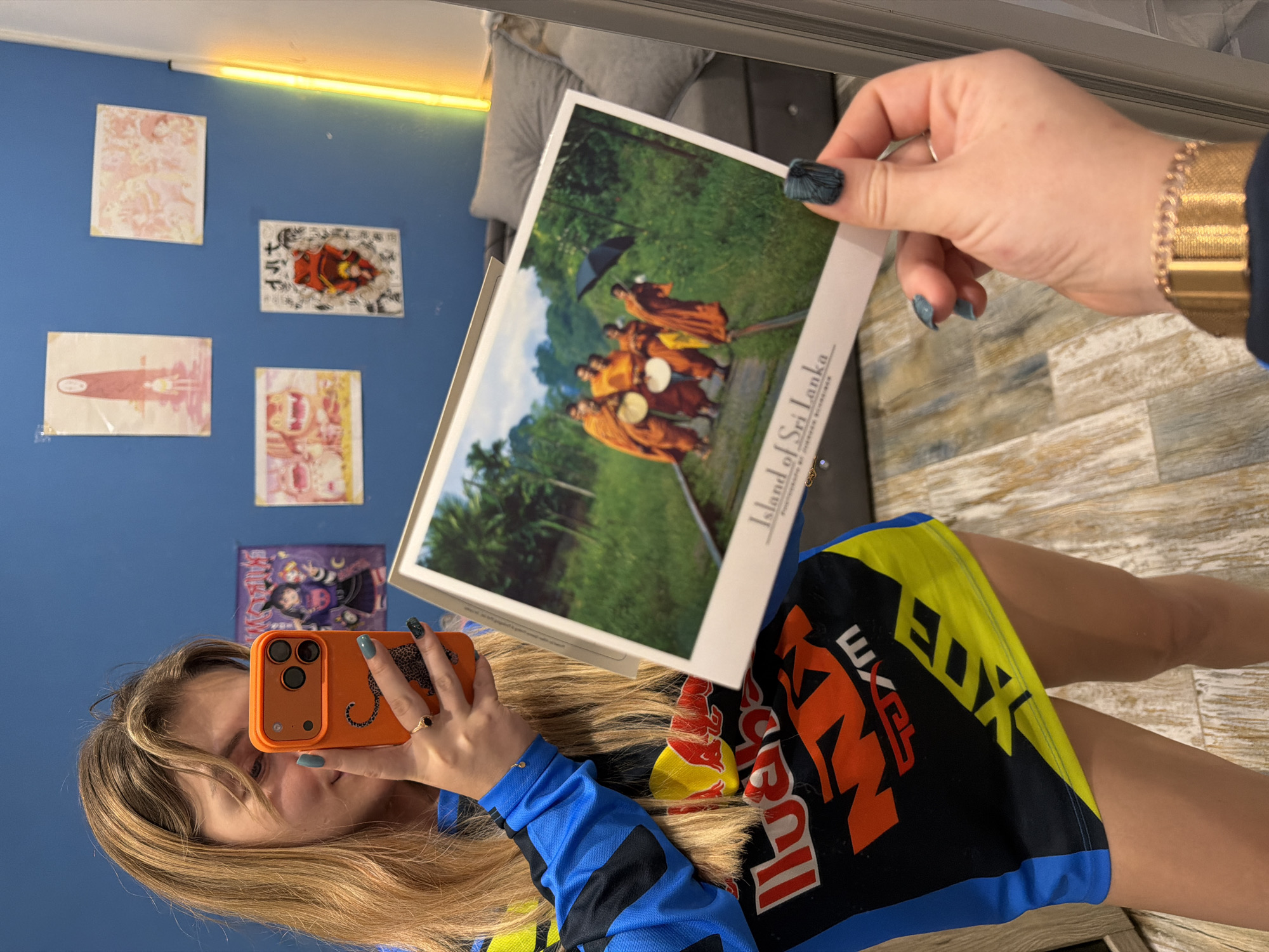 A person is holding up a postcard in front of a mirror, likely taking a photo of themselves in the reflection. The postcard depicts a group of monks in orange robes, with the words "Island of Sri Lanka" visible on the side. The person holding the card has blue and black nail polish on their fingernails and a gold watch on their wrist. They are wearing a blue and black motocross jersey with orange and yellow accents.
The reflection in the mirror shows part of a room with a blue wall. There are various small posters or pictures hanging on the wall. There is a yellow light fixture near the top of the wall. Part of a grey couch or chair is also visible in the upper portion of the mirror's reflection.
Based on the GPS data, the location associated with the image is in Большая Шильна, Russia. However, this information is not directly visually verifiable in the image itself.