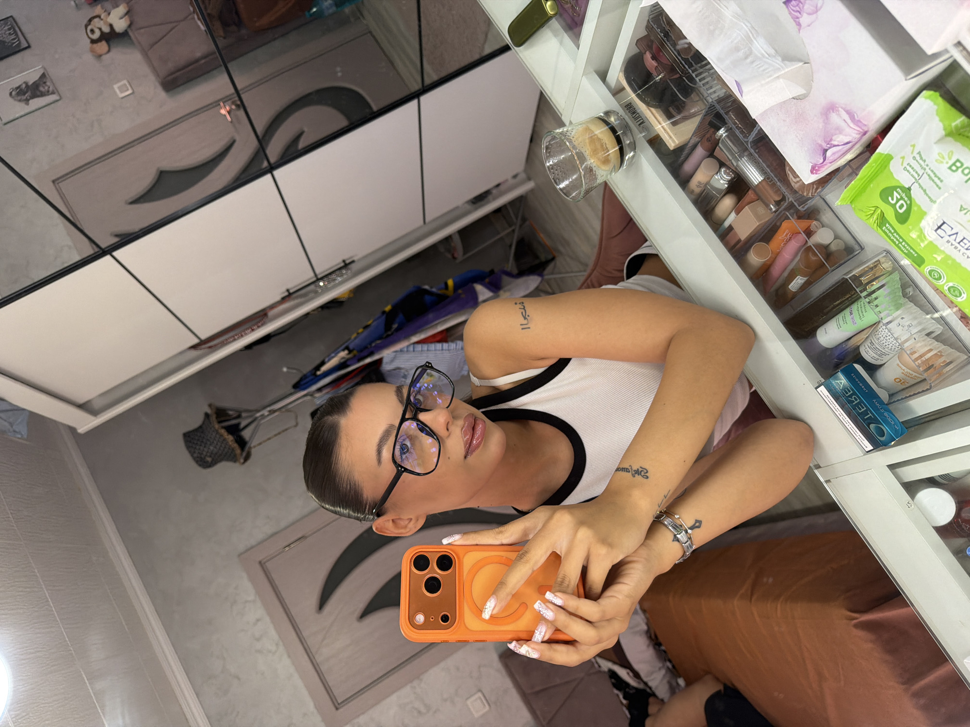 In a domestic setting, likely an apartment in Burgas, Bulgaria, a young woman is captured in a mirror selfie. She is positioned in the foreground, facing the camera, and holding an orange iPhone in her right hand. The woman is wearing a white tank top with black trim and has her hair pulled back into a sleek bun. She has on large black-rimmed glasses and her makeup is noticeable, with a focus on her lips. A tattoo is visible on her left forearm.
The background reveals a well-organized vanity area with numerous makeup products neatly arranged in drawers. To the right, a glass of espresso sits on the surface, beside a tissue box and other cosmetics. Behind her, a large wardrobe with mirrored doors reflects the room, showing a bed and closet doors. The overall environment appears to be a personal dressing area within a residential space in Burgas.