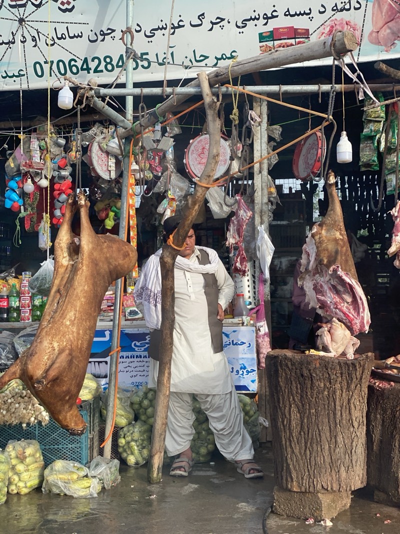 The image depicts a butcher shop in Mir Ahmad Kalay, Afghanistan. A man stands in the center of the shop, wearing traditional Afghan clothing. He has a scarf draped around his neck and a vest over his long-sleeved shirt. He stands in front of two large wooden poles supporting the shop's structure. Hanging on either side of him are carcasses of animals, possibly lambs or goats. To the left of the man, there is a stack of vegetables, including what appears to be zucchini, packaged in plastic bags. Behind the man, there is a sign with text written in Pashto or Dari. To the right, there are chopping blocks. The shop is open-air, with various items hanging from the ceiling, including light bulbs, decorations, and what appear to be small drums. The ground in front of the shop appears to be wet.