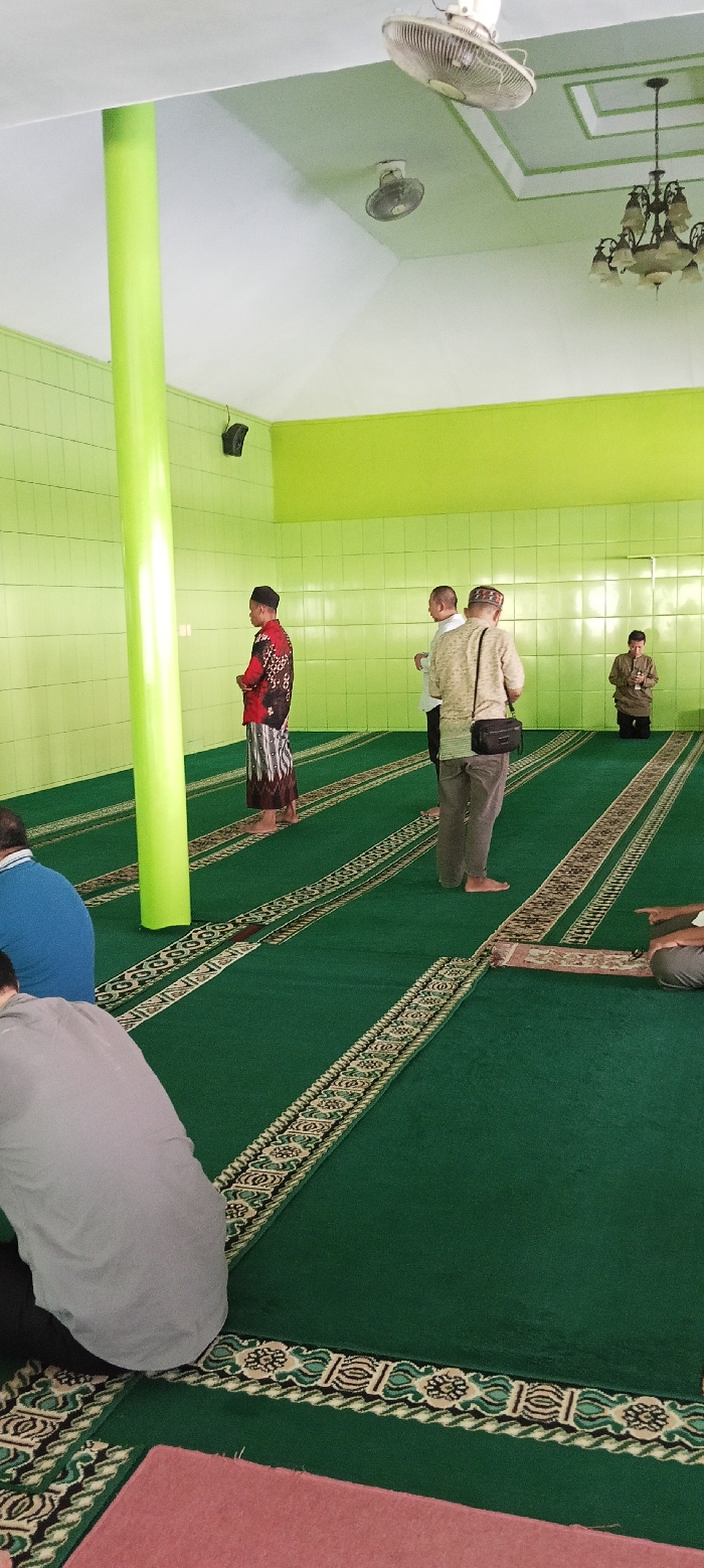 The interior of a mosque in Soreang, Indonesia, located at the coordinates -7.022, 107.525.  The mosque has a green tiled floor, with green carpet with intricate patterned borders.  There is a large pillar in the center of the room.  A ceiling fan is visible in the corner of the room.  Five men are in the room, four standing, one seated on the carpet.  Two men are standing in the center of the room, near the pillar.  A third man is standing by the wall in the distance.  A fourth man is kneeling in prayer next to the wall in the distance.  A fifth man is seated, turned toward the wall, on the green carpet in the foreground.  The men are wearing casual attire.  The room is illuminated by fluorescent lighting.  The scene depicts an interior space where people are engaged in prayer.