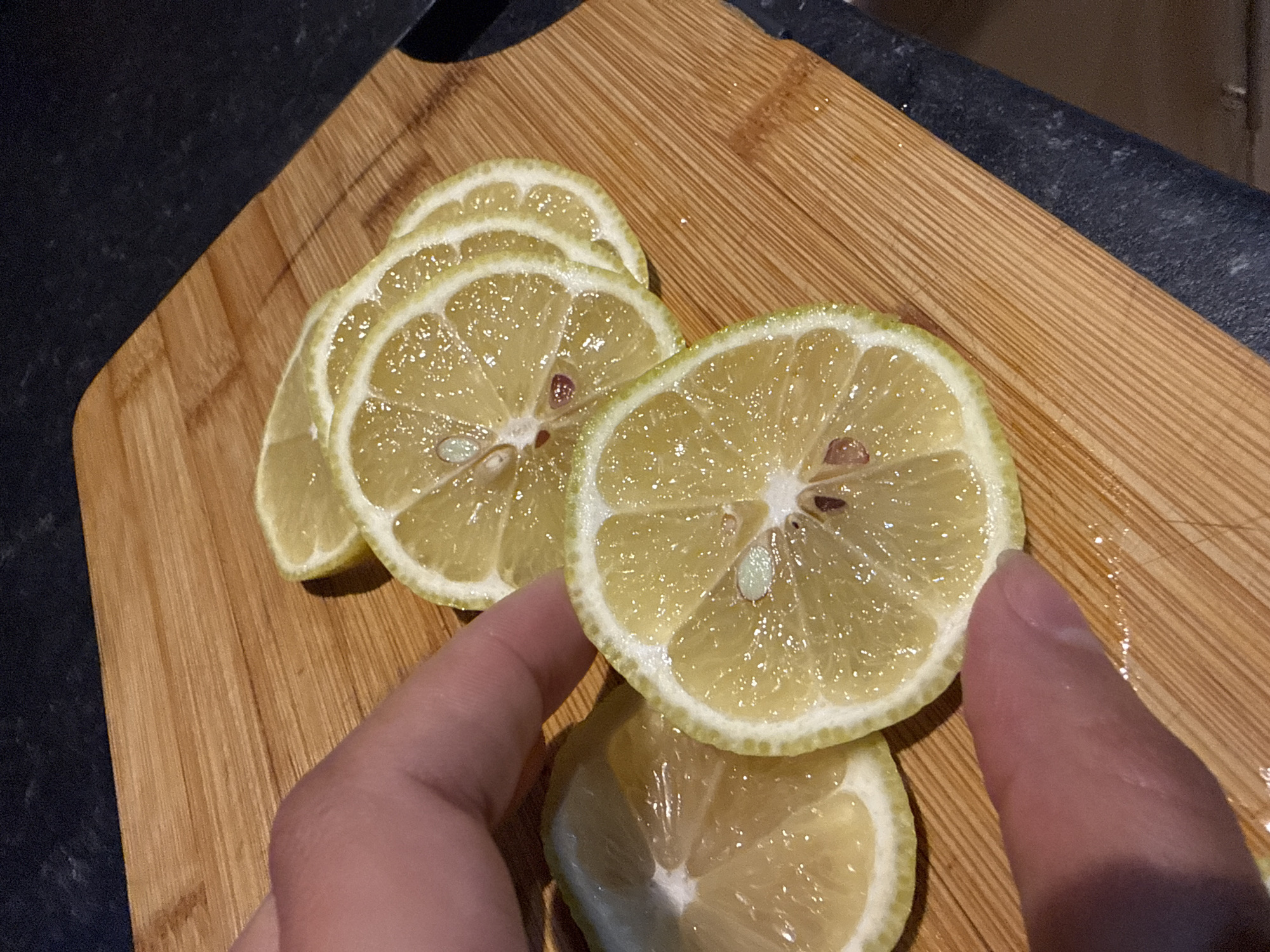 A high-angle, close-up shot shows a person's hand holding a lemon slice on a wooden cutting board. There are several other lemon slices on the board, arranged in a slightly overlapping manner. The lemons appear ripe, with a pale yellow flesh and a thin, light green rind. The hand is holding one of the slices, displaying it to the viewer. The cutting board has a distinct wood grain pattern with a light brown color. A dark surface, possibly a countertop, is visible in the background. There is no information to suggest the location is Great Yarmouth, United Kingdom.