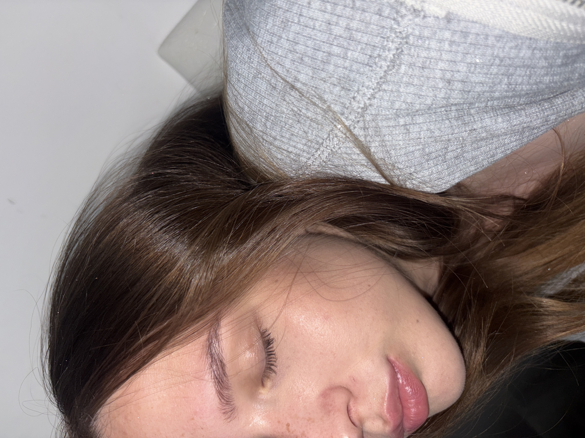 A close-up, eye-level shot shows the side profile of a young person with brown hair and fair skin, resting their head on a grey, ribbed garment. Their eyes are closed, and their lips are slightly parted. Freckles are visible on their cheek. The background is a plain, light-colored wall, with a white object positioned to the right of the person's head. There is no visible indication of the city of Chaikovsky, Russia, in this image.