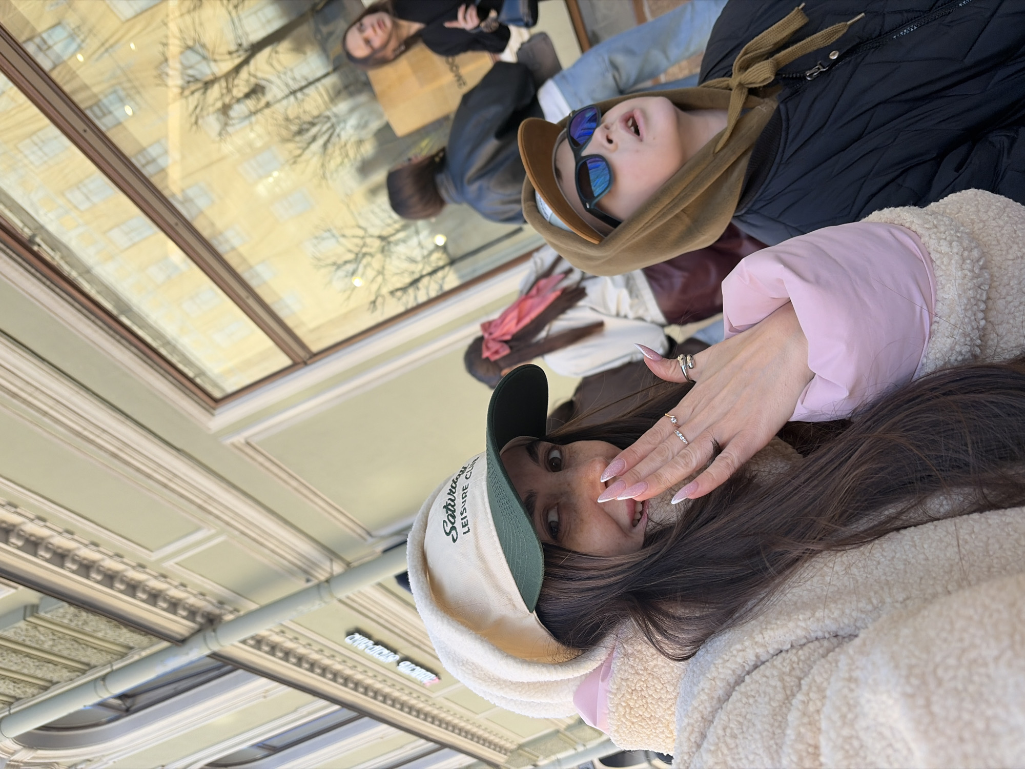 Two women are visible. One is wearing a beige sherpa jacket with pink accents, a Saturday Leisure Club hat, and has long brown hair. Her manicured hand, adorned with rings, partially covers her face. The second woman, positioned in the background, wears a dark puffy jacket over a brown hoodie, with sunglasses. Reflections in a window show additional figures, including another woman in a black top and blue jeans carrying a shopping bag. The reflections also reveal a building facade and bare tree branches. The location is a building in Saint Petersburg, Russia, evidenced by signage and architectural details on the ceiling. The women appear to be standing near a window in a commercial area.