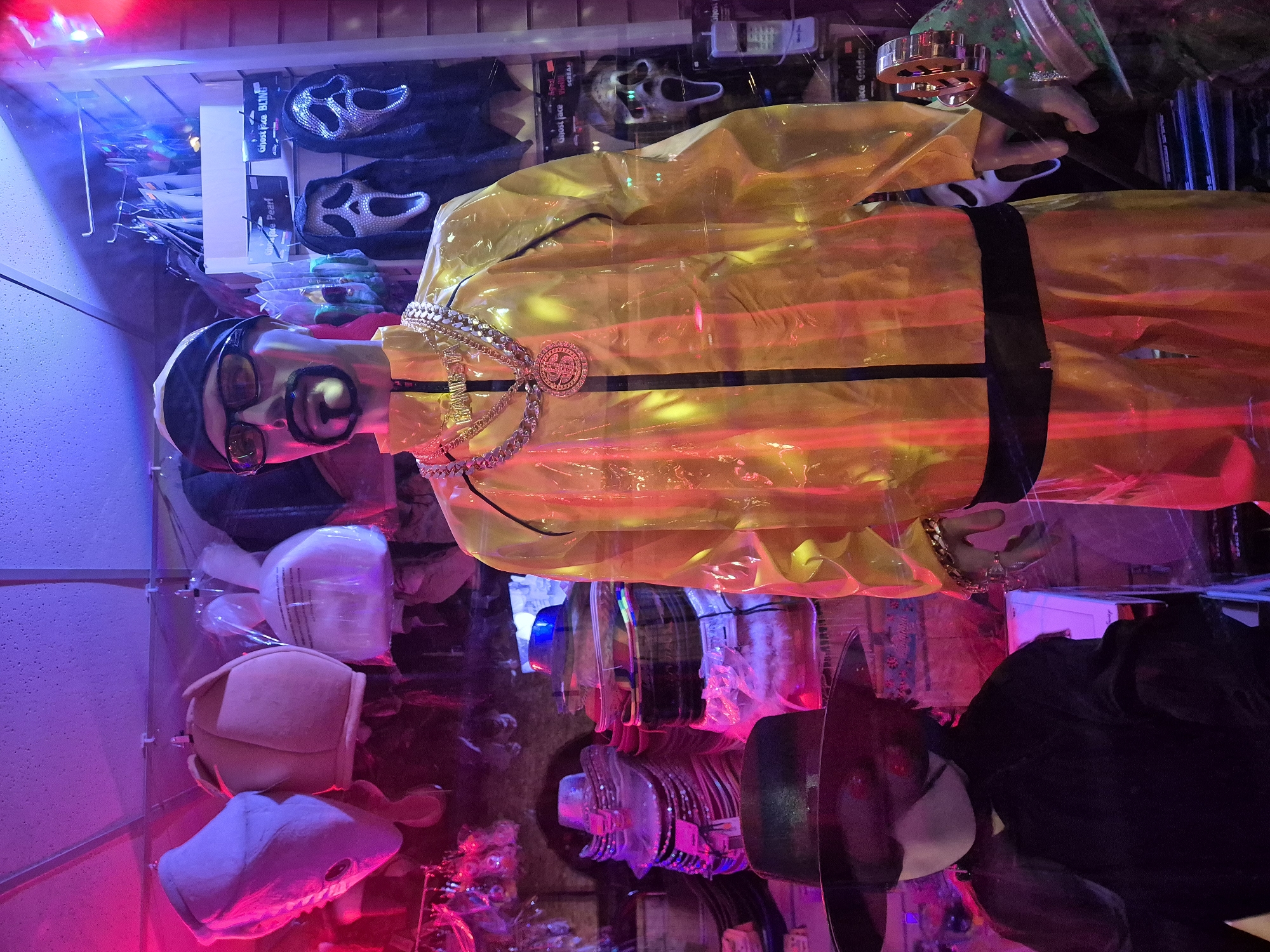 This image shows a mannequin dressed in a yellow raincoat and accessorized with gold chains, glasses, and a mustache. It appears to be displayed in a shop window in Cardiff, United Kingdom. Other items in the window include masks and hats. The scene is illuminated with pink and blue lighting.