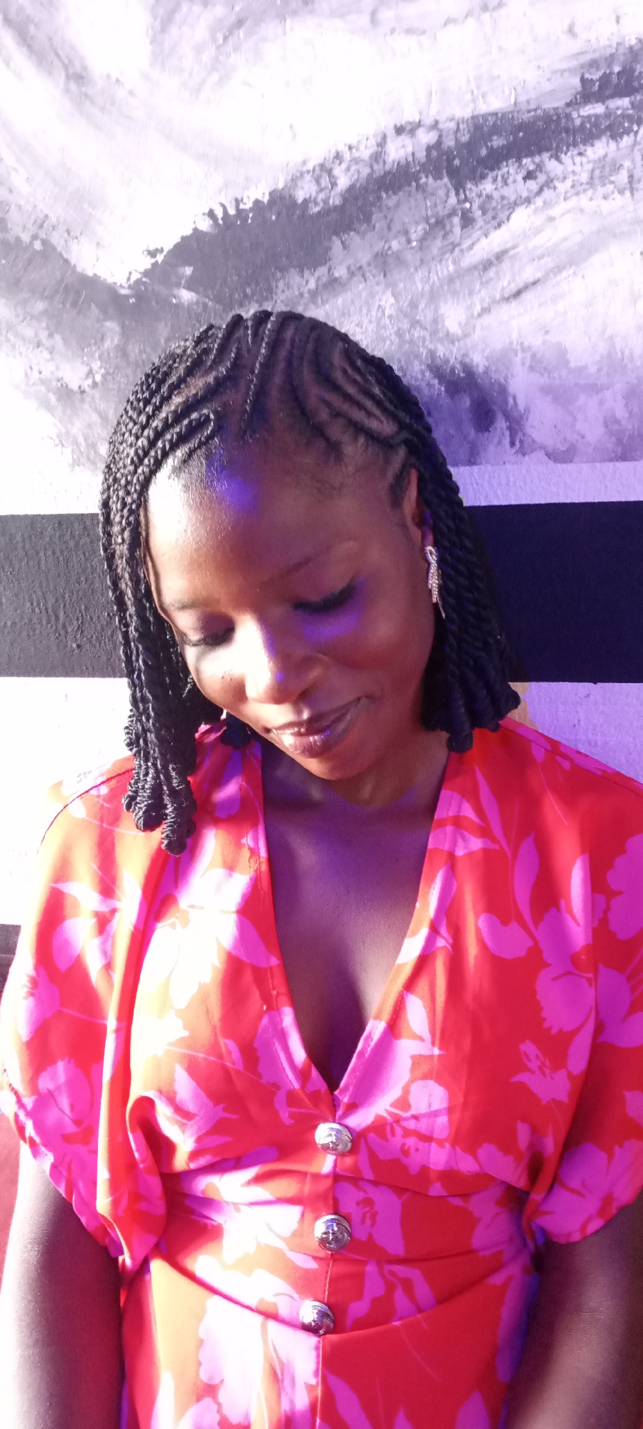 The image depicts a woman with braided hair, wearing a floral print top with buttons, posed in front of a patterned wall. The wall behind her has horizontal stripes of white/grey and black, and a textured pattern in the white/grey areas. The background is lit with a purple hue. No other details are available to confidently state the location is in Jos, Nigeria.