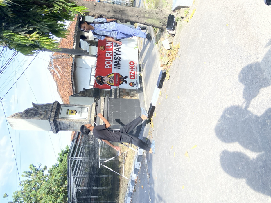 The image shows two young men walking on a paved street in front of a building with a sign that reads "POLRI UNT MAS YARA." The man on the left is wearing a black t-shirt, black pants, and black shoes. He is walking away from the camera. The man on the right is wearing a blue and white checkered shirt, blue pants, and black shoes. He is walking towards the camera. The sign is red and white with a cartoon image of a police officer in the top left corner. The sign is attached to a brick building with a stone column in front of it. There is a green tree on the right side of the image. The location of the image is in Purwakarta, Indonesia, based on the provided coordinates. The shadows of the two young men are clearly visible on the ground. The location is a street in Purwakarta, Indonesia. The environment is an urban setting.