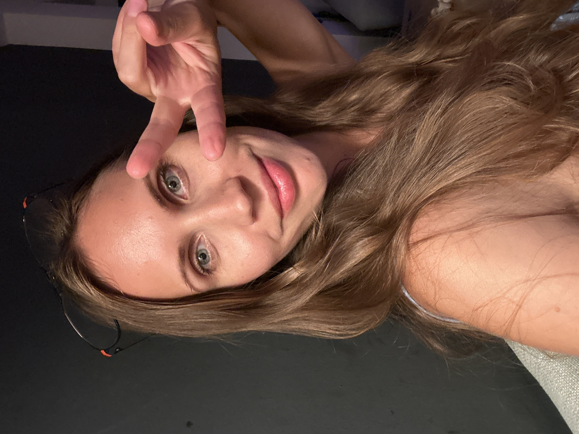 A young woman with long, light brown hair, wearing glasses perched on her head, is taking a selfie indoors. She is looking at the camera with a slight smile and holding up two fingers in a peace sign gesture. Her skin appears tanned, and she is wearing a light-colored top. The background is dark and appears to be a wall. The scene is likely a personal space, such as a home or hotel room, and is not indicative of a specific public location in Fethiye, Turkey.
