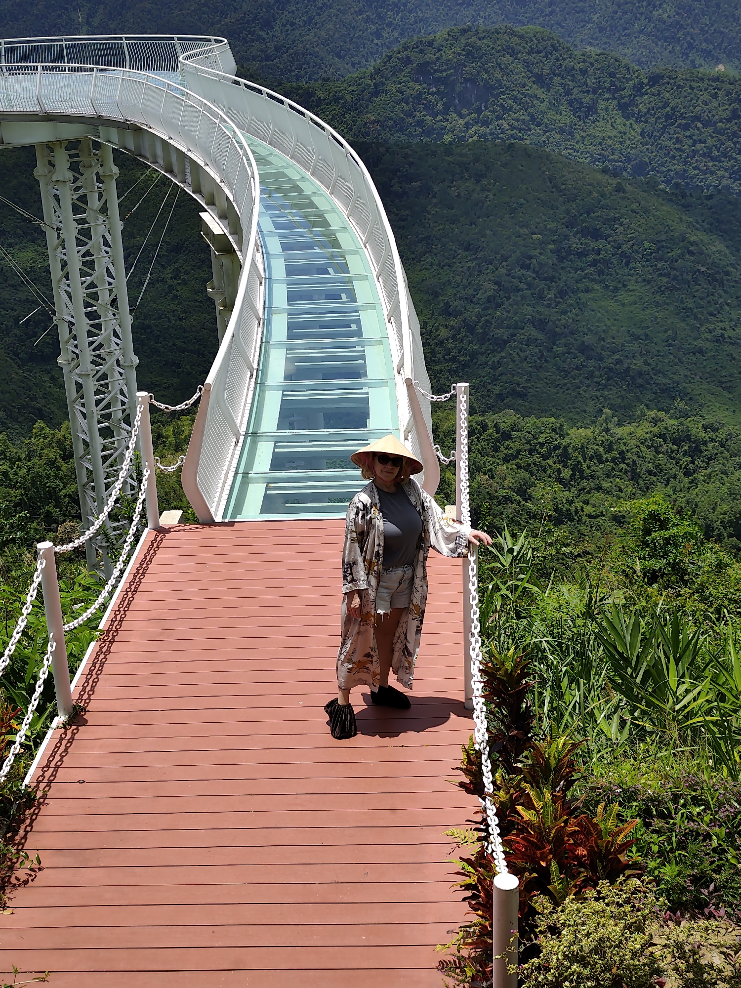 A woman stands on a wooden walkway next to a transparent glass bridge that curves upwards. The walkway is made of reddish-brown planks, and white metal posts with chains connect to it, forming a barrier. The woman is wearing a straw hat, sunglasses, a patterned robe, a grey t-shirt, and denim shorts. She is holding onto the chain barrier with her right hand. The glass bridge, supported by white metal structures, offers a view of lush, green mountains in the background. The environment is tropical, with various plants and trees visible.