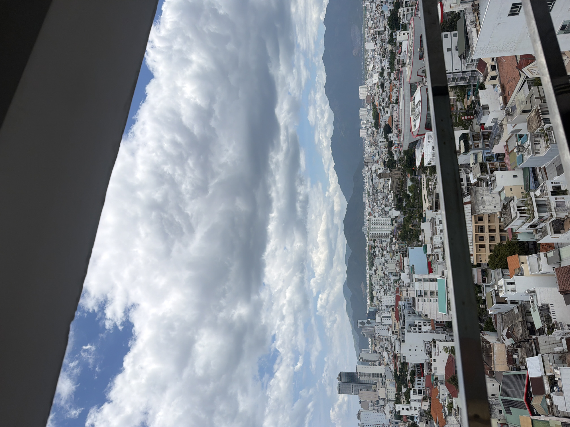 The image shows an elevated view of the city of Nha Trang, Vietnam, on a partly cloudy day. In the foreground, a metal railing is visible, suggesting the photo was taken from a balcony or elevated platform. Beyond the railing, a dense urban landscape unfolds, characterized by numerous buildings of varying heights and architectural styles, predominantly white and light-colored. Some buildings appear to be residential apartments, while others might be commercial structures.
In the mid-ground, a prominent set of modern buildings with distinctive curved, white and red structures stands out. Adjacent to these is a tall, older-style building with a steeple, likely a church. Further into the cityscape, rows of lower-rise buildings with tiled roofs are visible, interspersed with pockets of greenery. In the distance, a range of dark, undulating mountains forms the horizon, partially obscured by clouds.
The sky is a mix of bright blue patches and large, textured clouds, some of which appear heavy and gray, indicating potential for rain. The overall impression is that of a bustling coastal city nestled against a backdrop of natural mountainous terrain, under a dynamic sky. The scene depicts an urban environment with a clear sense of depth and perspective, leading the eye from the immediate foreground to the distant mountains. No people are visible in the image.